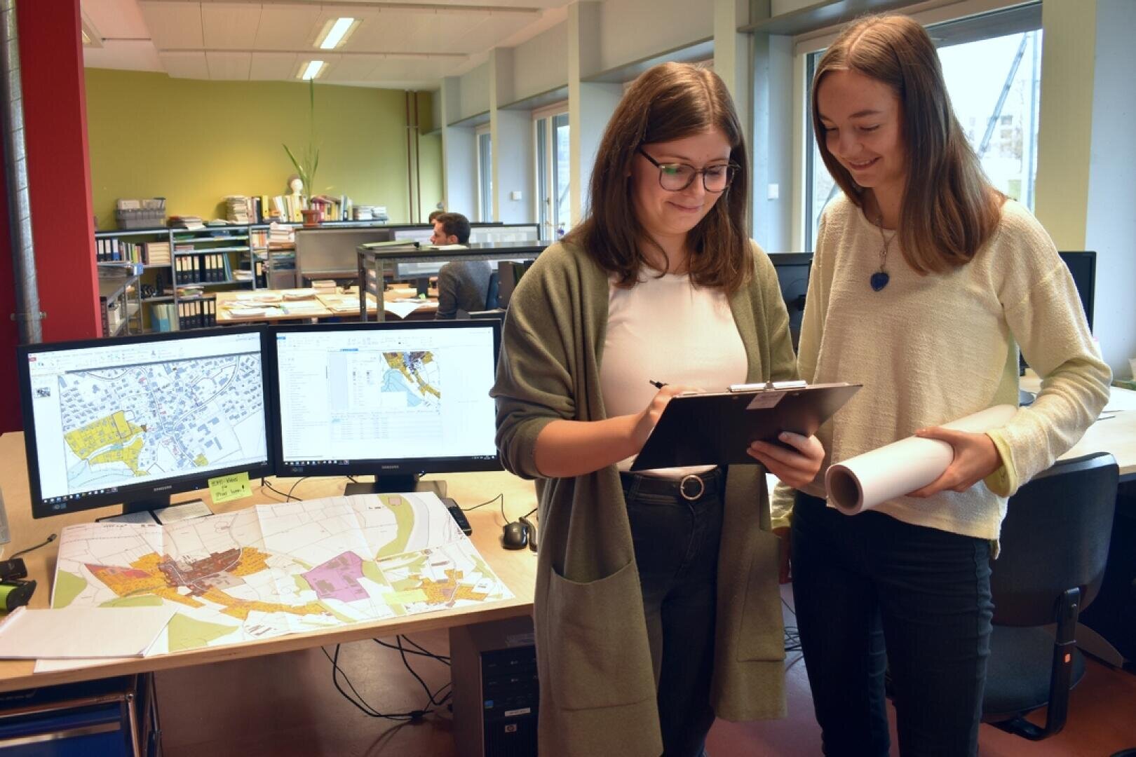 Deux femmes se tiennent dans un bureau, l'une tenant une planchette à pince et l'autre des papiers enroulés. Derrière elles, deux moniteurs affichent des cartes et un bureau est couvert d'impressions de cartes. D'autres personnes travaillent à l'arrière-plan.