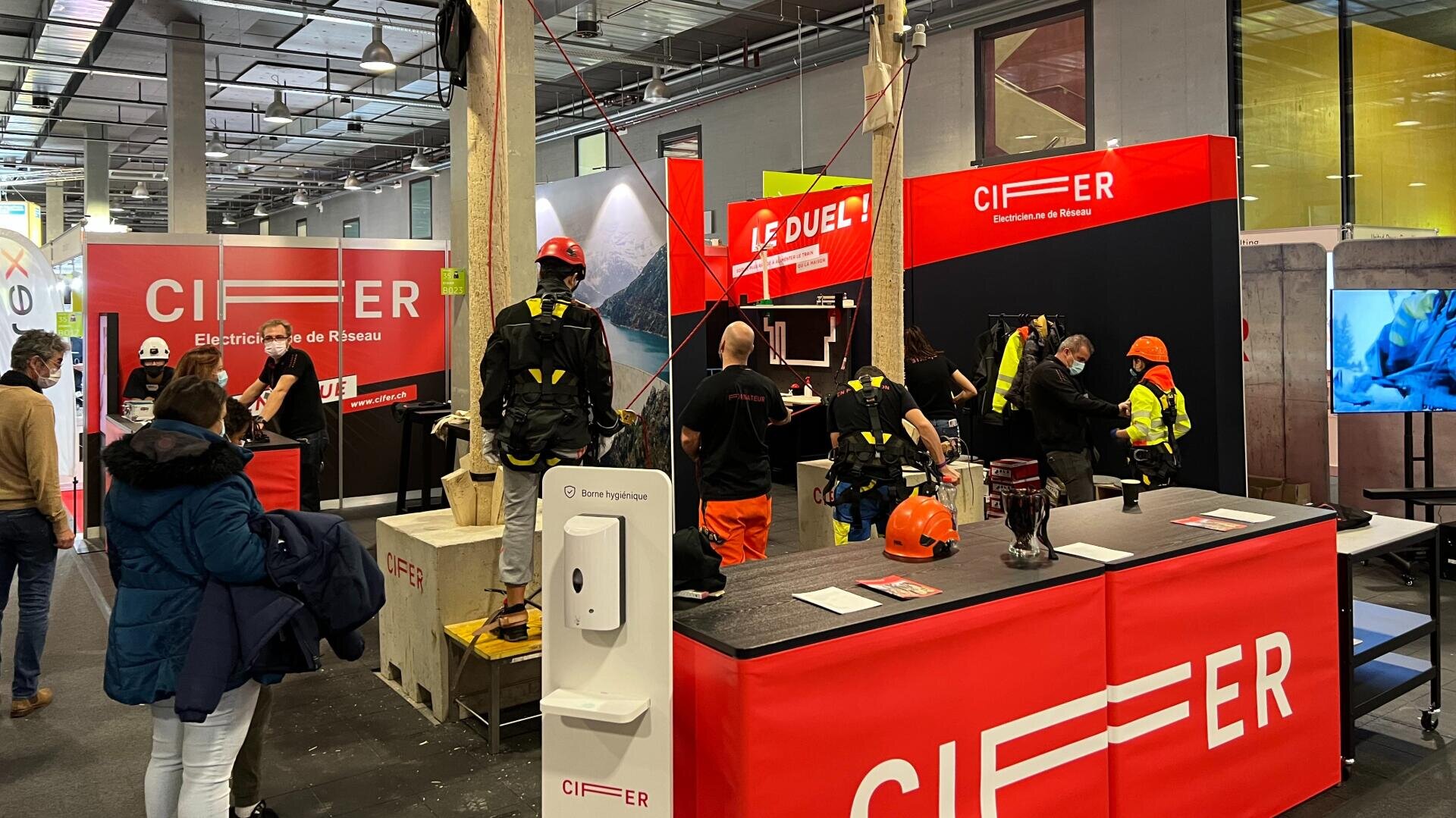 Un stand de CIFER sur un salon professionnel, avec des personnes en tenue de chantier, un comptoir rouge, des équipements de sécurité et un panneau "LE DUEL". Les visiteurs observent les démonstrations et les expositions sous un éclairage intérieur intense.