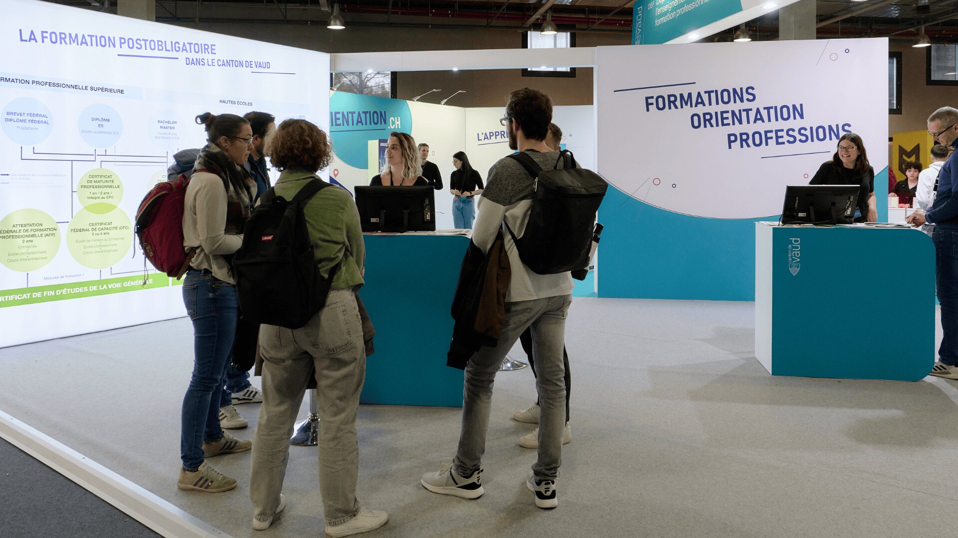 Un groupe de jeunes avec des sacs à dos se tient devant les stands d'information d'un salon de l'éducation et discute avec le personnel. De grands panneaux derrière les stands affichent des informations sur l'enseignement postsecondaire et l'orientation professionnelle.
