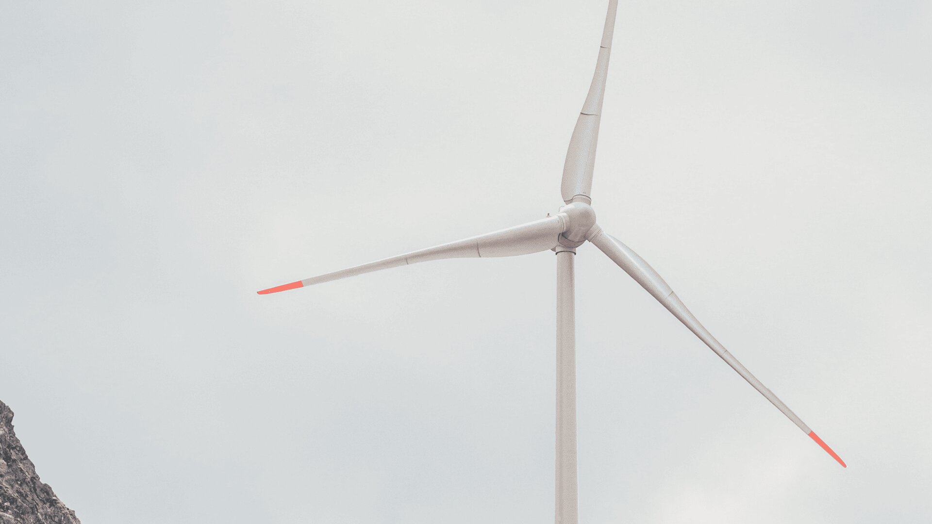 Vue rapprochée d'une éolienne à trois pales, chacune de couleur rouge, sur un ciel nuageux et une petite colline rocheuse dans le coin.