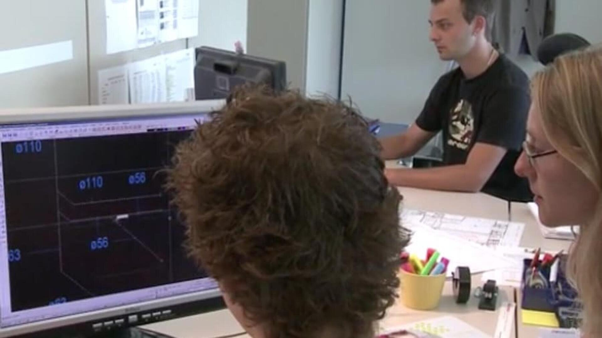 Deux personnes regardent un écran d'ordinateur affichant des dessins techniques, tandis qu'une autre personne travaille à un bureau à l'arrière-plan. L'espace de travail comporte des papiers, des fournitures de bureau et un grand écran sur lequel sont affichés des plans.