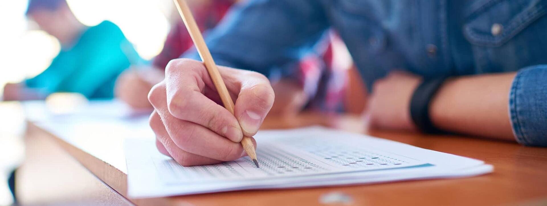 Gros plan d'une main tenant un crayon et remplissant une feuille de réponses à choix multiples à un bureau en bois, avec d'autres personnes travaillant à l'arrière-plan.