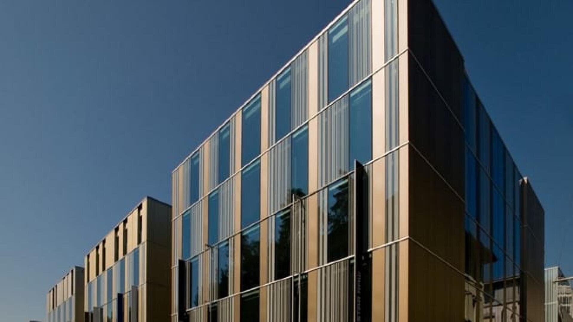 Bâtiment moderne avec de grandes baies vitrées et des panneaux métalliques verticaux, sur fond de ciel bleu clair. La structure présente des lignes épurées et une conception architecturale contemporaine.