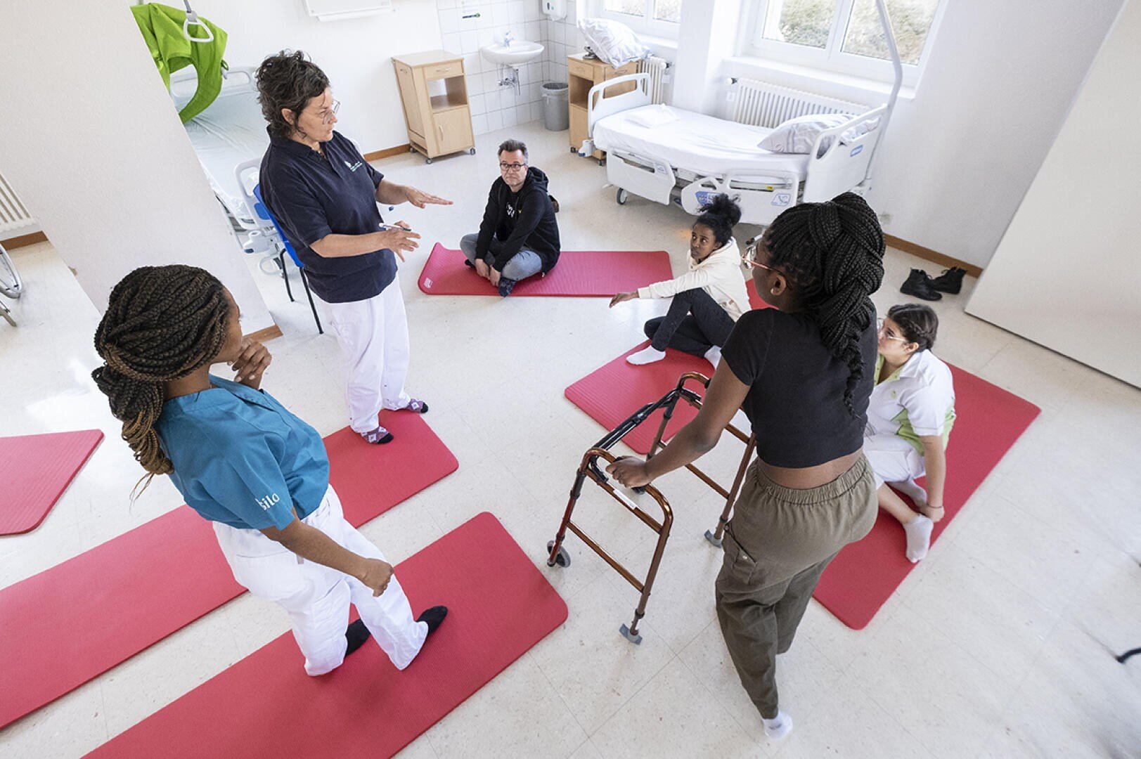 Un groupe de personnes participe à une séance de kinésithérapie ou de rééducation sur des tapis rouges dans une salle médicale lumineuse, avec une personne utilisant un déambulateur et un instructeur donnant des instructions.