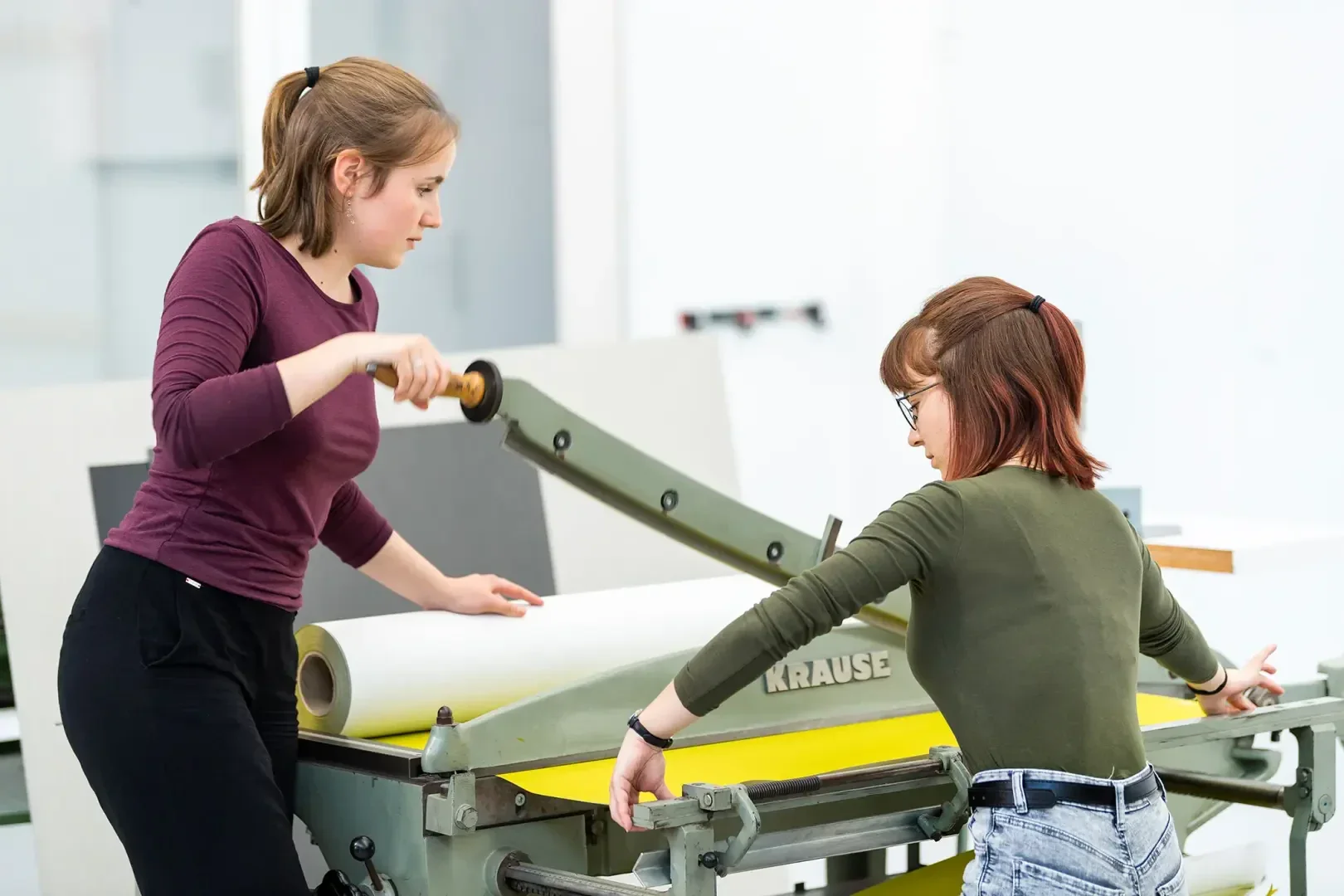 Deux femmes font fonctionner une grande presse à imprimer. L'une tourne la poignée tandis que l'autre ajuste le papier sur le plateau de la presse. Toutes deux se concentrent sur leur tâche dans un espace de travail moderne et lumineux.