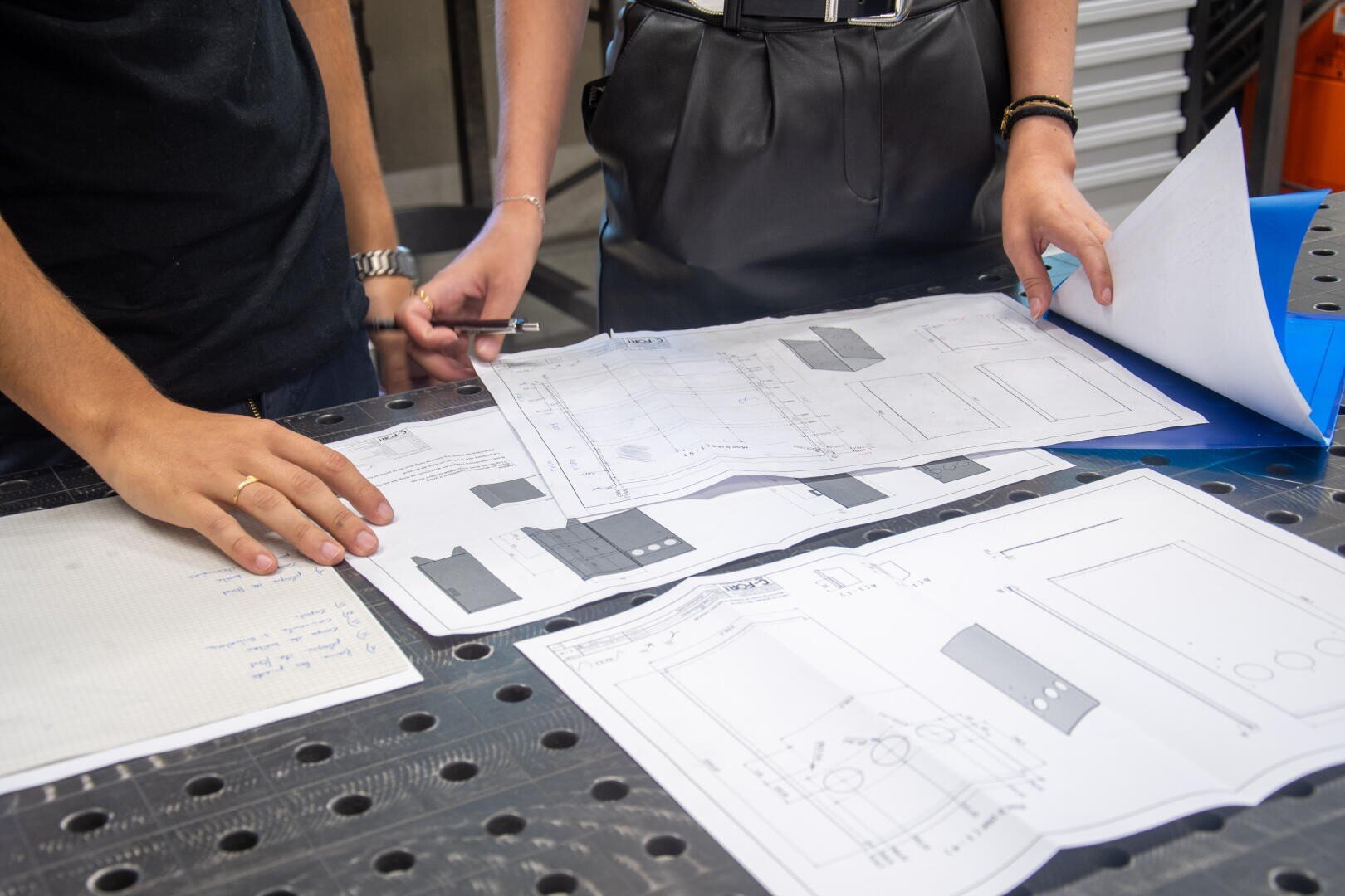 Deux personnes examinent des dessins techniques et des plans répartis sur un établi ; l'une tient un stylo et l'autre un dossier contenant d'autres documents. Seuls leurs mains et leurs torses sont visibles.