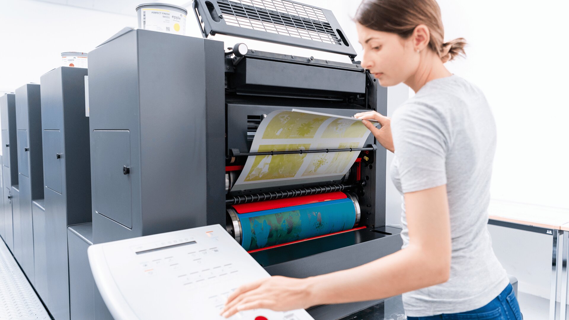 Une femme fait fonctionner une grande presse industrielle, ajustant une feuille imprimée à l'intérieur de la machine tout en se tenant près d'un panneau de commande dans un espace de travail moderne et lumineux.