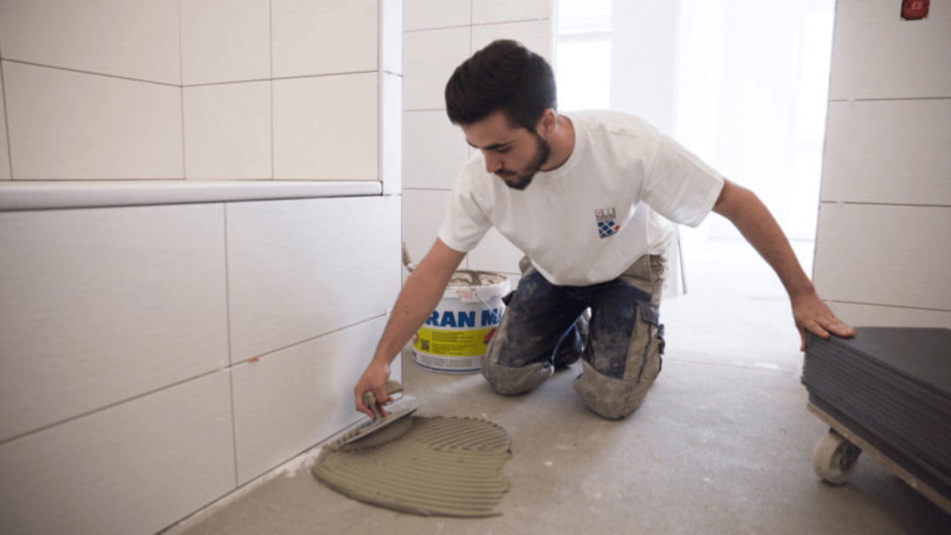 Un homme est agenouillé sur le sol, étalant de la colle sur le sol à l'aide d'une truelle dentelée lors de la pose de carreaux sur le mur d'une salle de bains. Des feuilles de carrelage et un seau se trouvent à proximité.