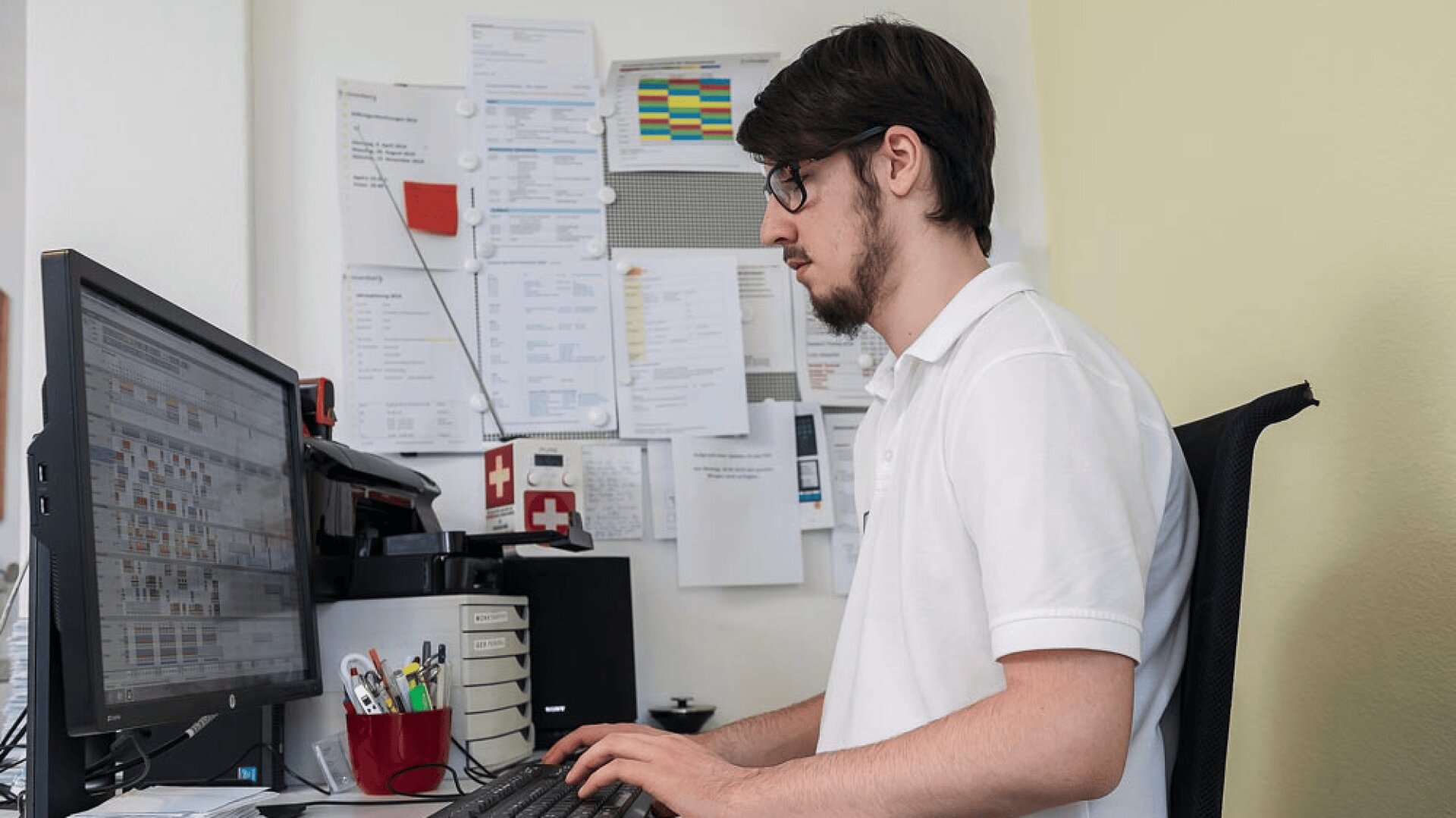 Un homme portant des lunettes et un polo blanc est assis à un bureau et tape sur un clavier. Il fait face à un écran d'ordinateur sur lequel s'affichent des tableaux et des documents. Des papiers et des notes sont accrochés au mur derrière lui.