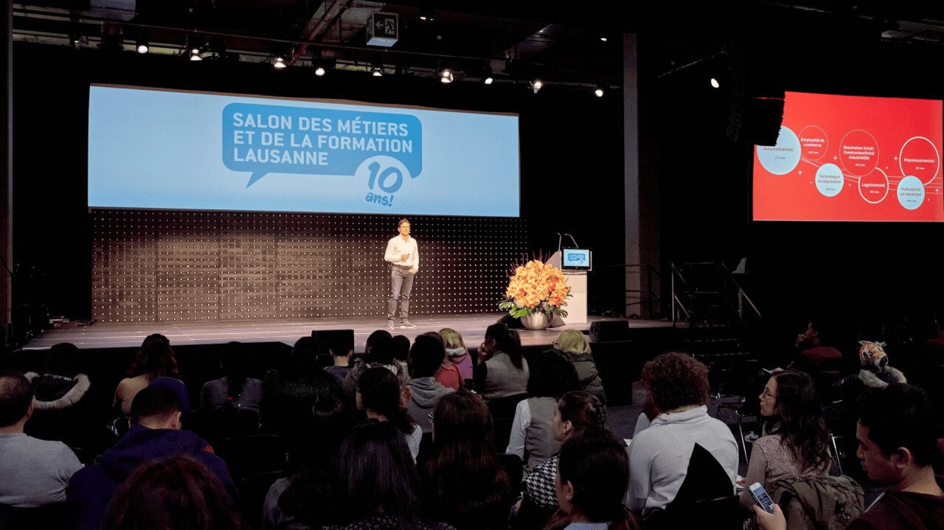 Un orateur se tient sur la scène du Salon des Métiers et de la Formation de Lausanne et s'adresse au public. Un grand écran affiche le nom et l'anniversaire de l'événement, tandis qu'un autre écran montre un diagramme.