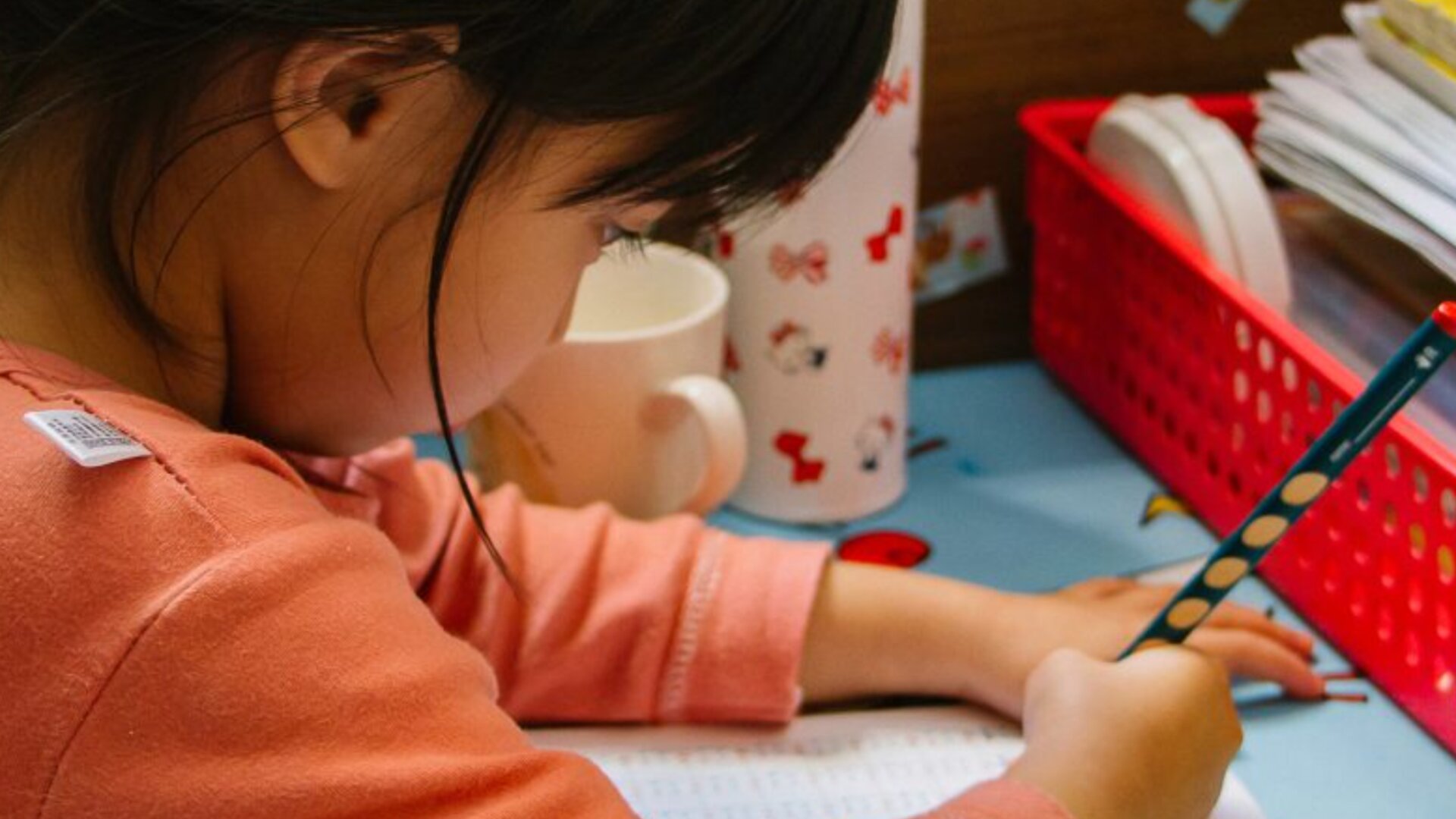 Un jeune enfant aux cheveux noirs écrit au crayon dans un cahier d'exercices à un bureau, entouré d'une tasse, de papiers et d'un panier rouge.