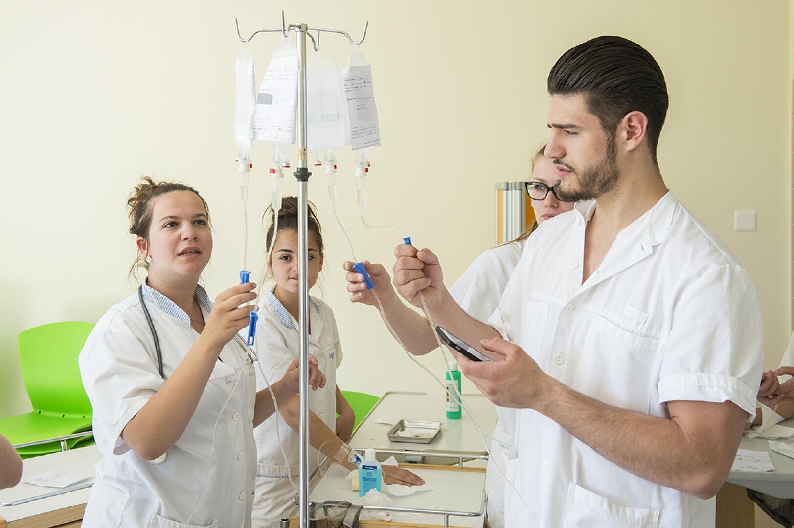 Quatre étudiants en médecine en blouse blanche travaillent ensemble à la préparation de solutés, examinent le matériel et prennent des notes dans une salle de classe bien éclairée, avec des chaises vertes en arrière-plan.