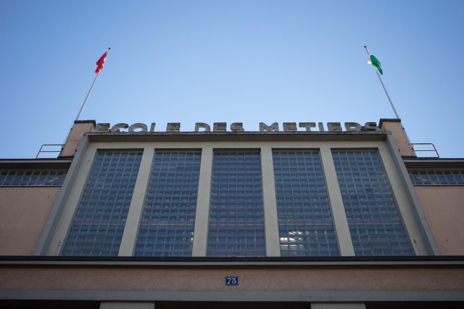 La façade d'un bâtiment avec de grandes baies vitrées et l'inscription Ecole des Métiers en haut, flanquée de deux mâts, l'un avec un drapeau rouge et l'autre avec un drapeau vert, sous un ciel bleu limpide.