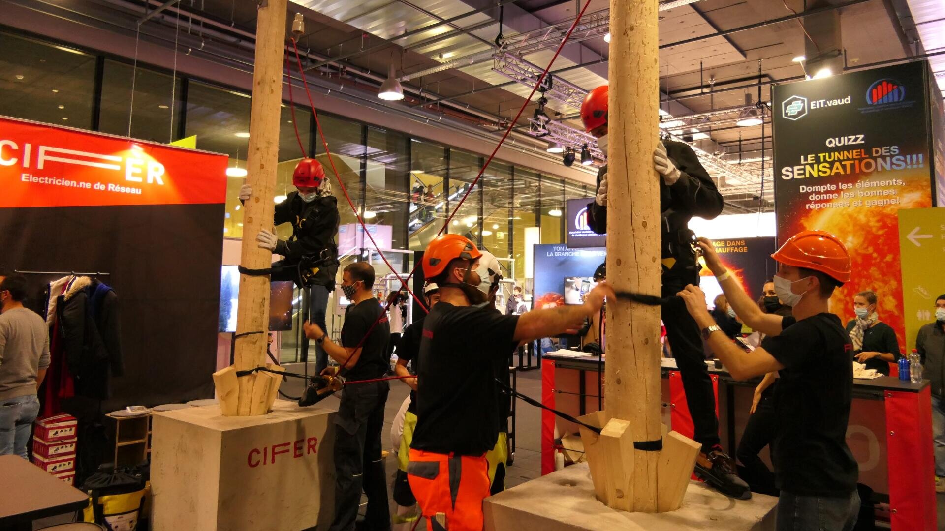 Des personnes portant des équipements de sécurité et des casques aident deux individus à grimper sur des poteaux en bois lors d'un événement ou d'une exposition en intérieur. Des stands, des bannières et des spectateurs sont visibles à l'arrière-plan.