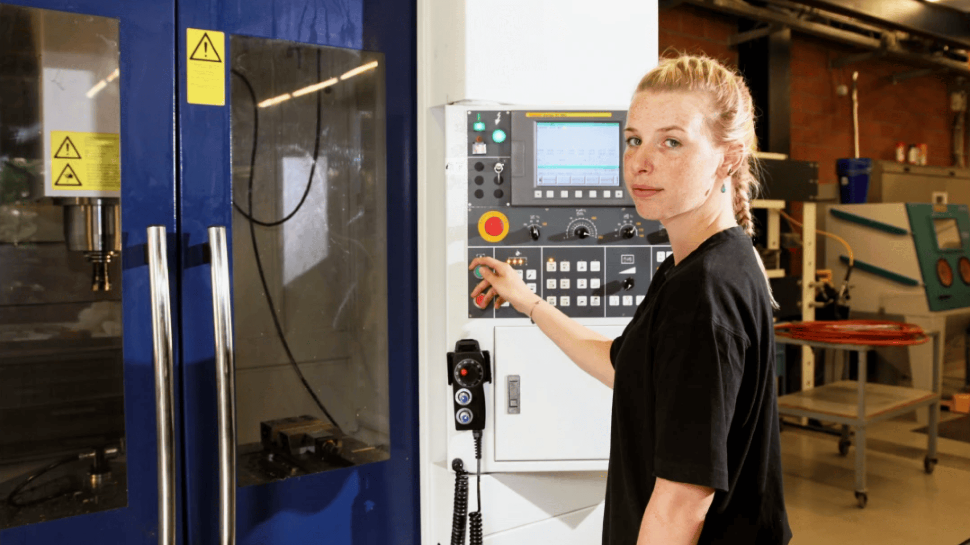 Une femme aux cheveux blonds attachés manipule un panneau de commande sur une machine industrielle dans une usine, en regardant vers la caméra.