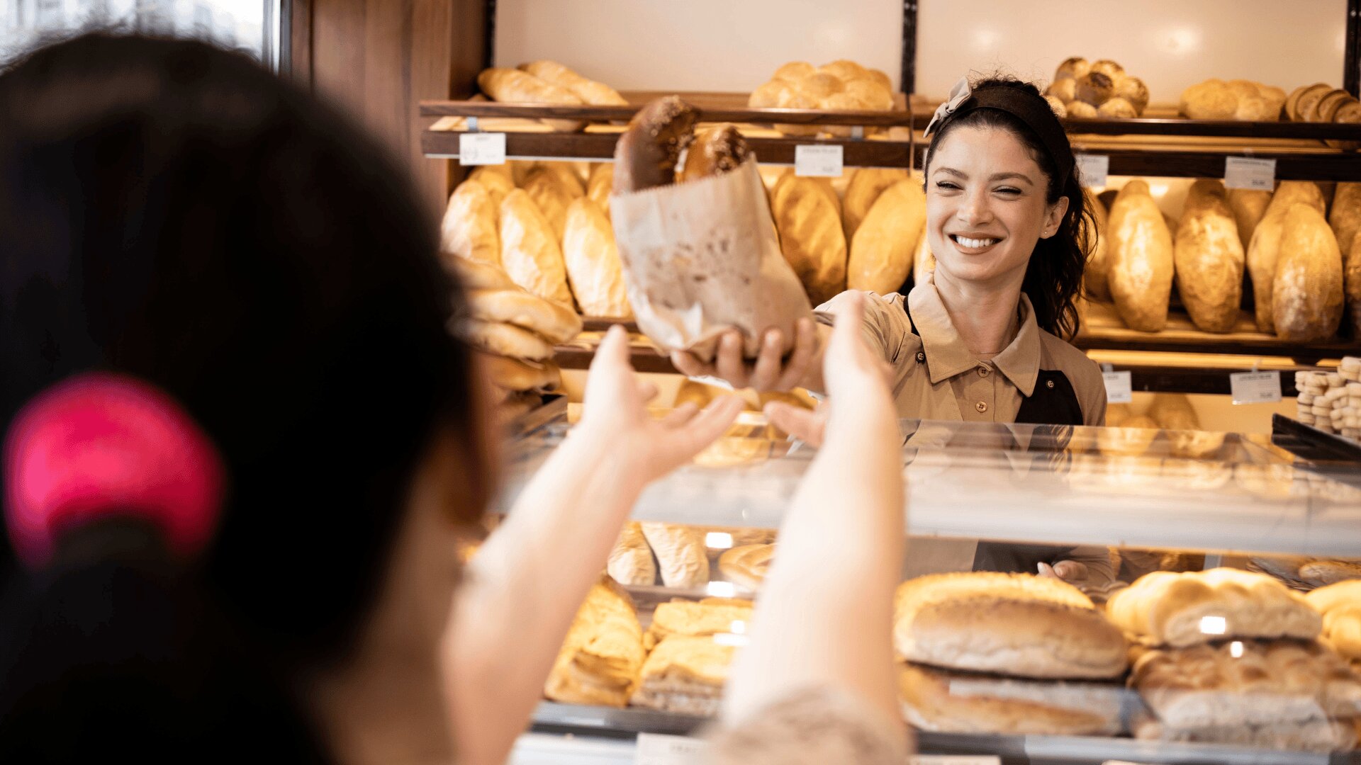 Un boulanger souriant tend une pâtisserie emballée à un client à travers un présentoir en verre rempli de pain et de produits de boulangerie, avec des étagères de pains à l'arrière-plan.