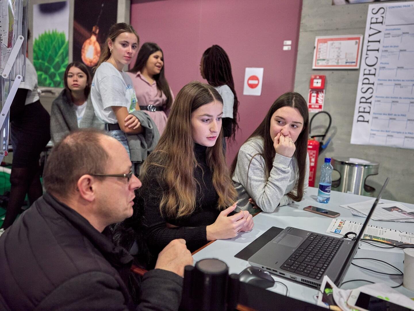 Un groupe de personnes réunies autour d'une table avec des ordinateurs portables et des documents ; deux jeunes femmes sont assises et concentrées sur l'ordinateur, tandis que d'autres se tiennent à proximité et observent attentivement dans un environnement intérieur.