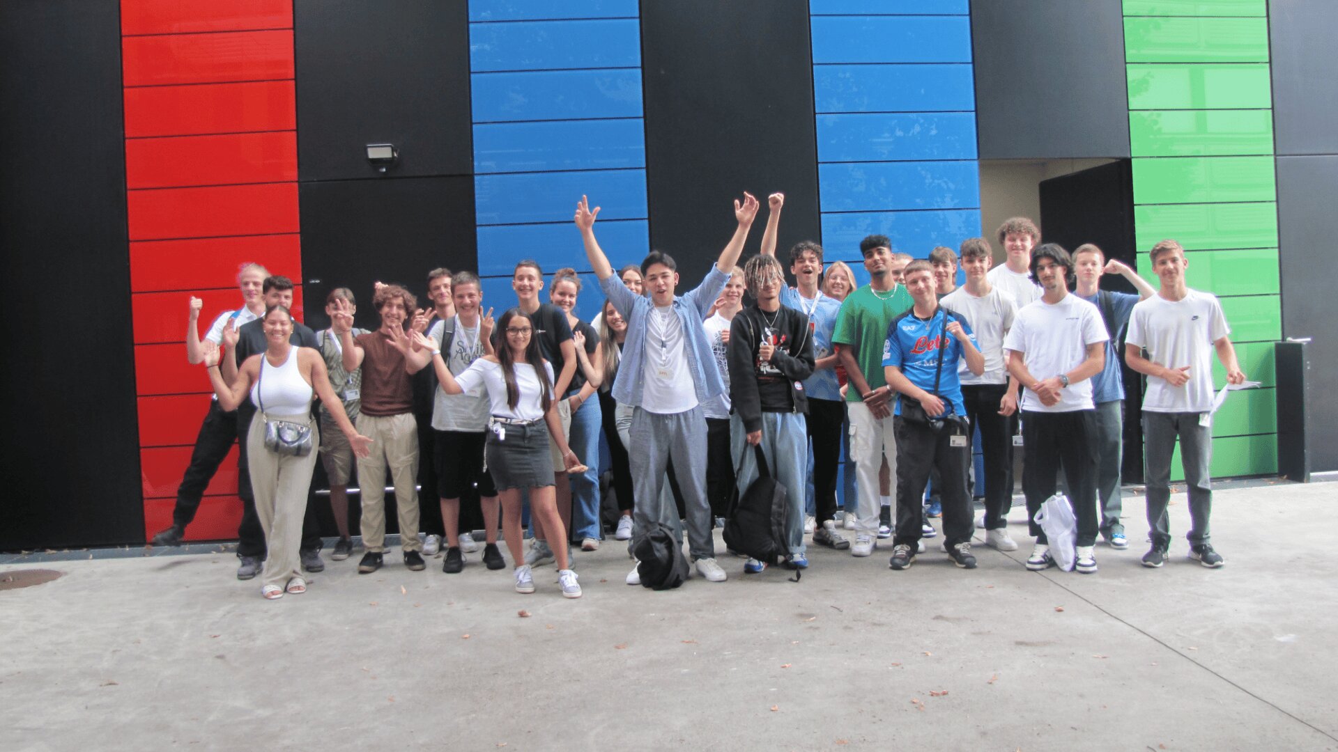 Un grand groupe de jeunes pose et sourit avec enthousiasme devant un bâtiment moderne aux panneaux rouges, bleus, verts et noirs. Certains ont les mains levées, des sacs à dos et des appareils photo sont visibles.