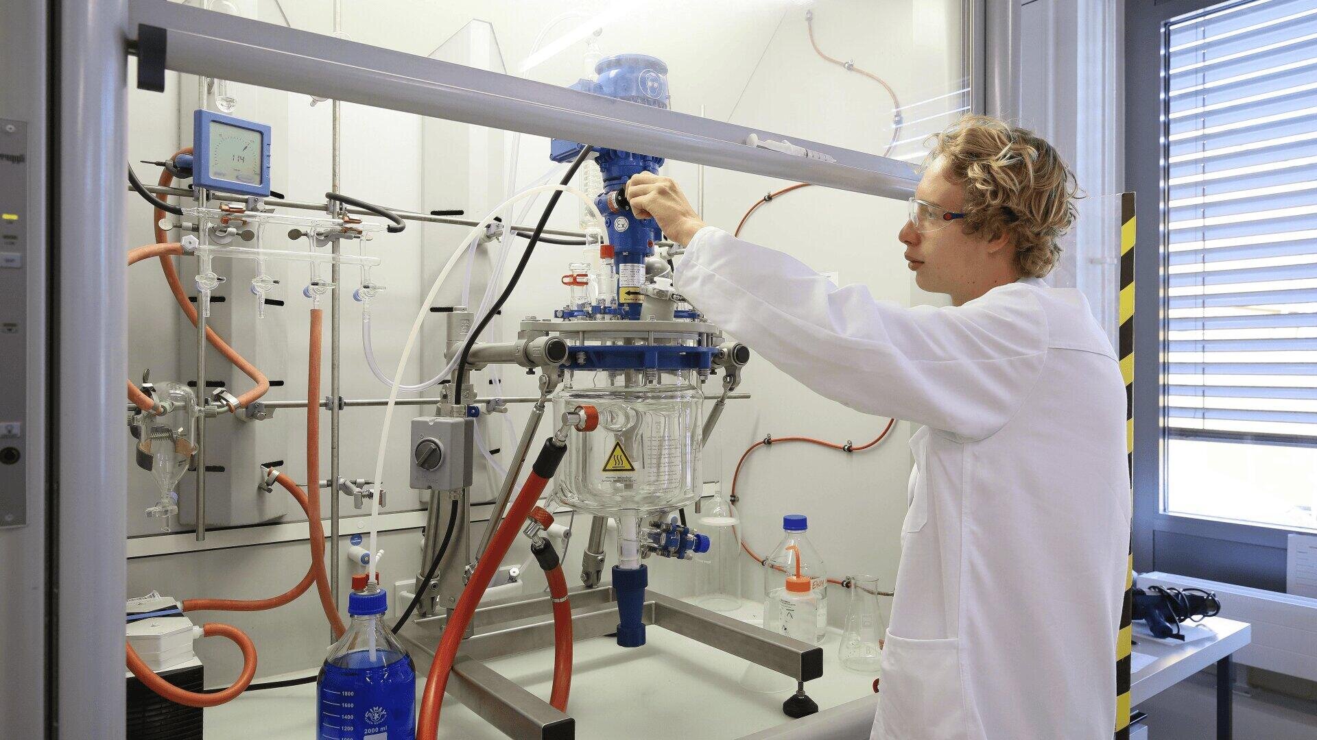 Une personne vêtue d'une blouse blanche et de lunettes de protection manipule un équipement scientifique dans un laboratoire, réglant une vanne sur une grande cuve de réacteur en verre entourée de tubes et d'instruments.