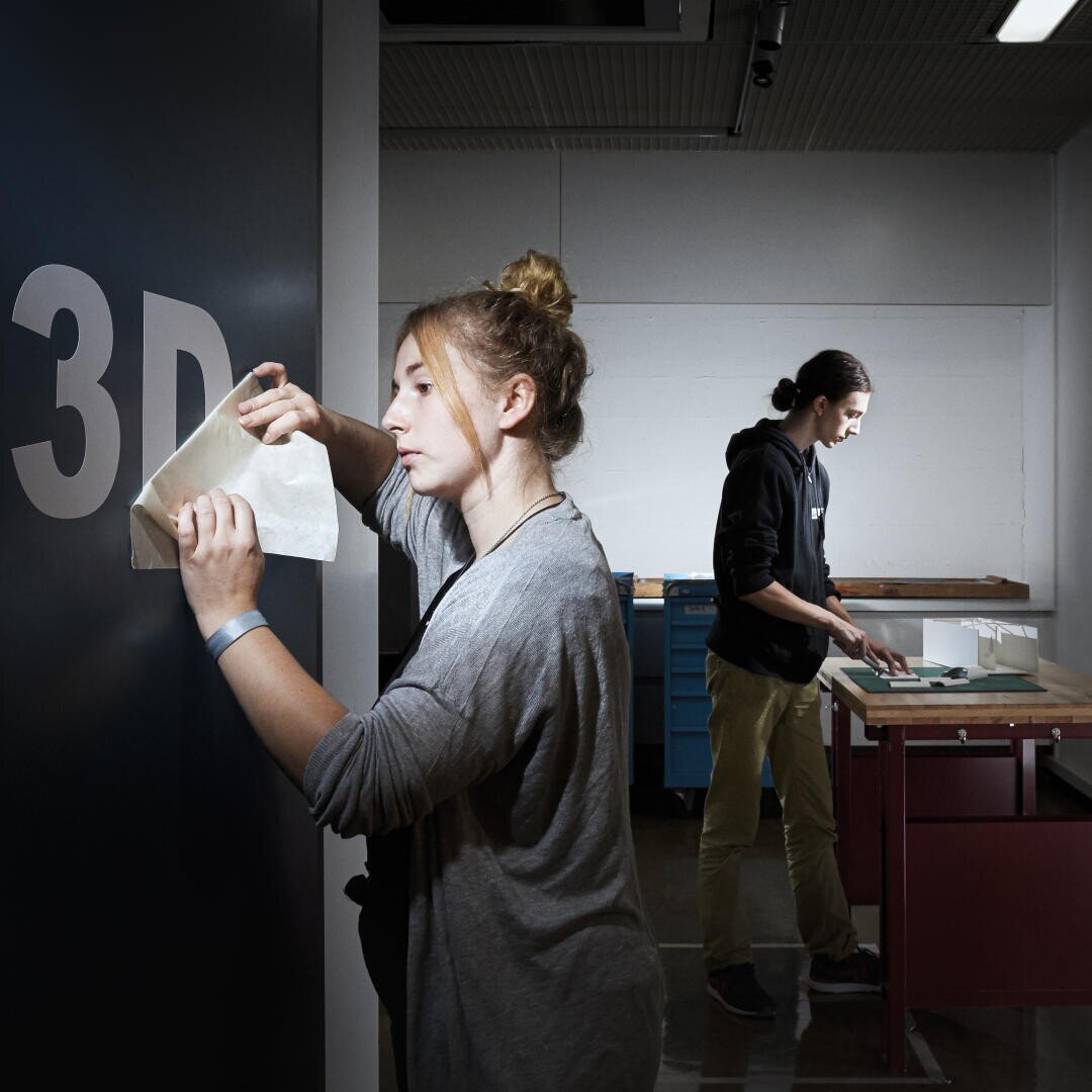 Une femme applique un panneau 3D sur un mur tandis qu'un homme travaille à une table à l'arrière-plan d'un atelier ou d'un makerspace bien éclairé.