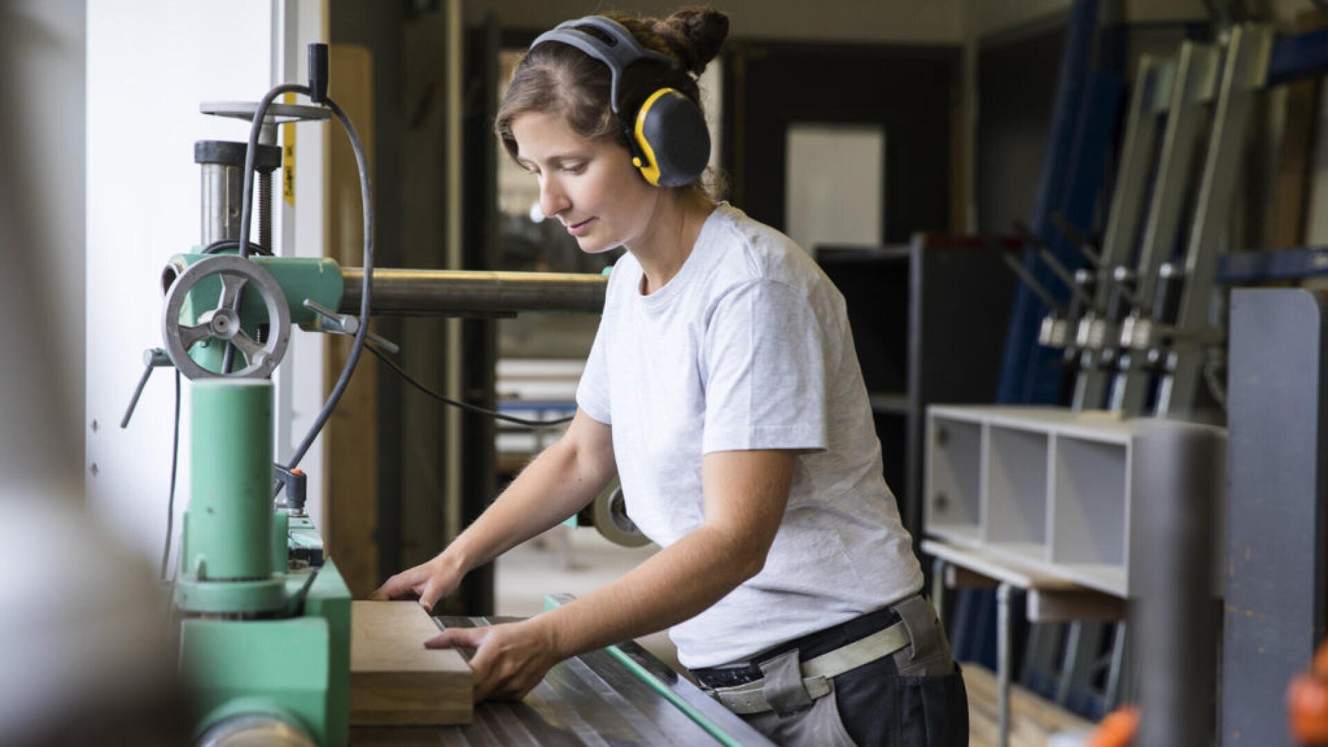Une femme portant des protections auditives et un t-shirt blanc utilise une machine à bois pour couper une pièce de bois dans un atelier. Divers outils et équipements sont visibles à l'arrière-plan.
