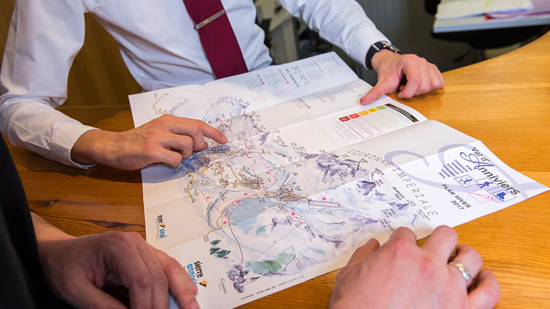 Deux personnes sont assises à une table en bois et examinent une carte détaillée d'une station de ski, l'une d'entre elles pointant du doigt un endroit précis sur la carte. Les deux personnes portent des vêtements d'affaires et semblent discuter des détails de la carte.