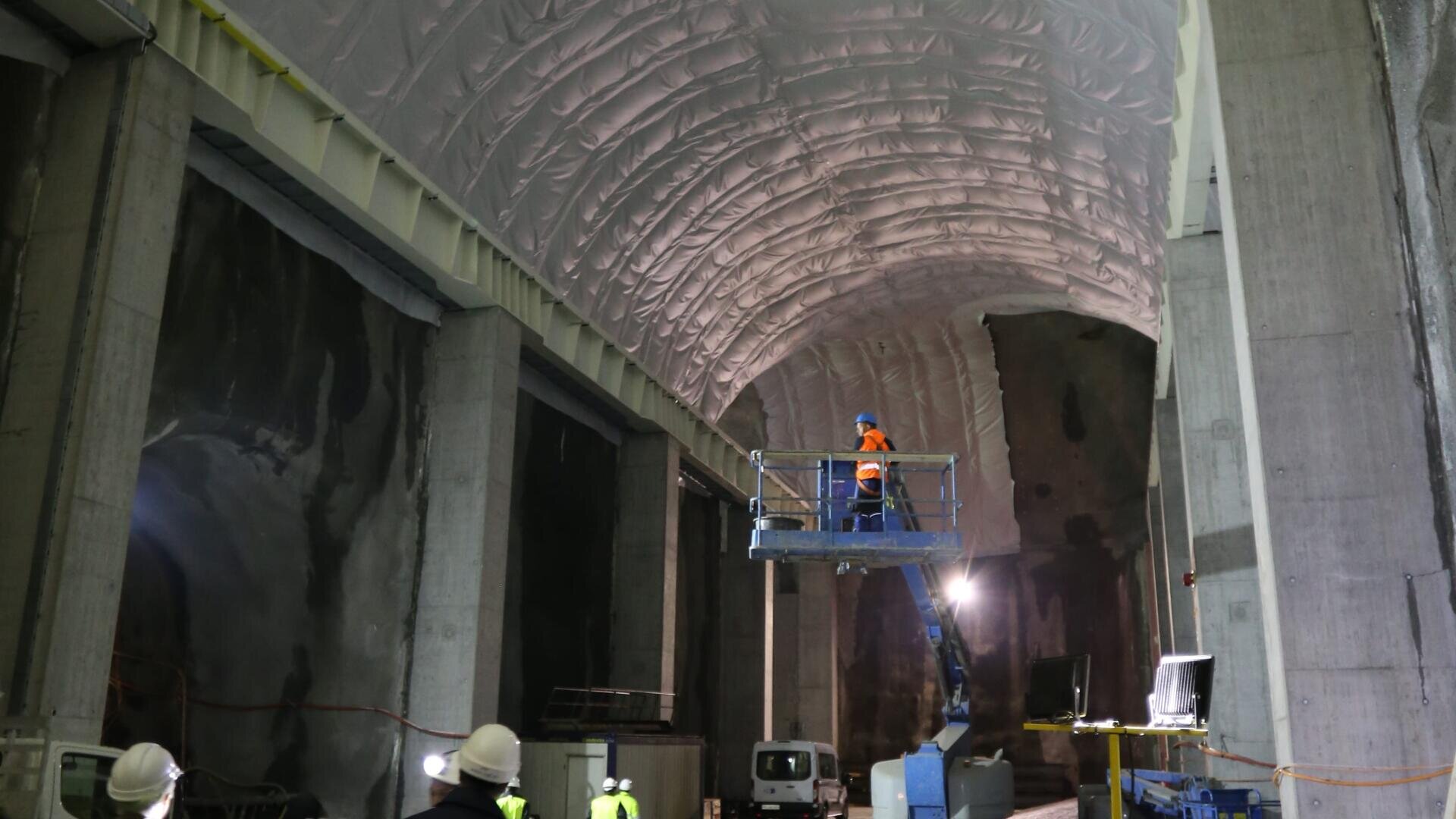 Des travailleurs en tenue de sécurité actionnent un ascenseur sous le plafond voûté et isolé d'un grand tunnel partiellement construit ou d'une installation souterraine dotée de murs en béton et d'équipements de construction.