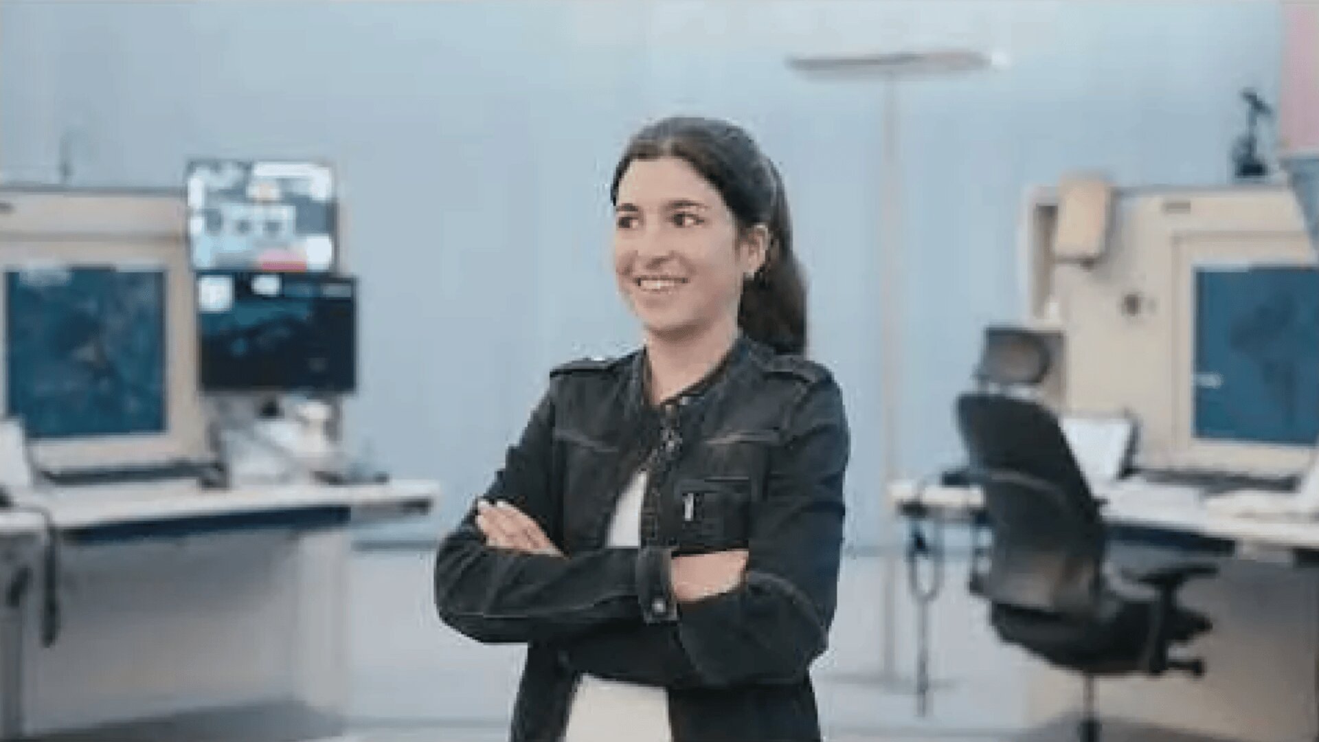 Une femme aux longs cheveux noirs, vêtue d'une veste noire et d'une chemise blanche, se tient debout, les bras croisés et sourit dans un bureau moderne avec des écrans d'ordinateur et des chaises pivotantes en arrière-plan.