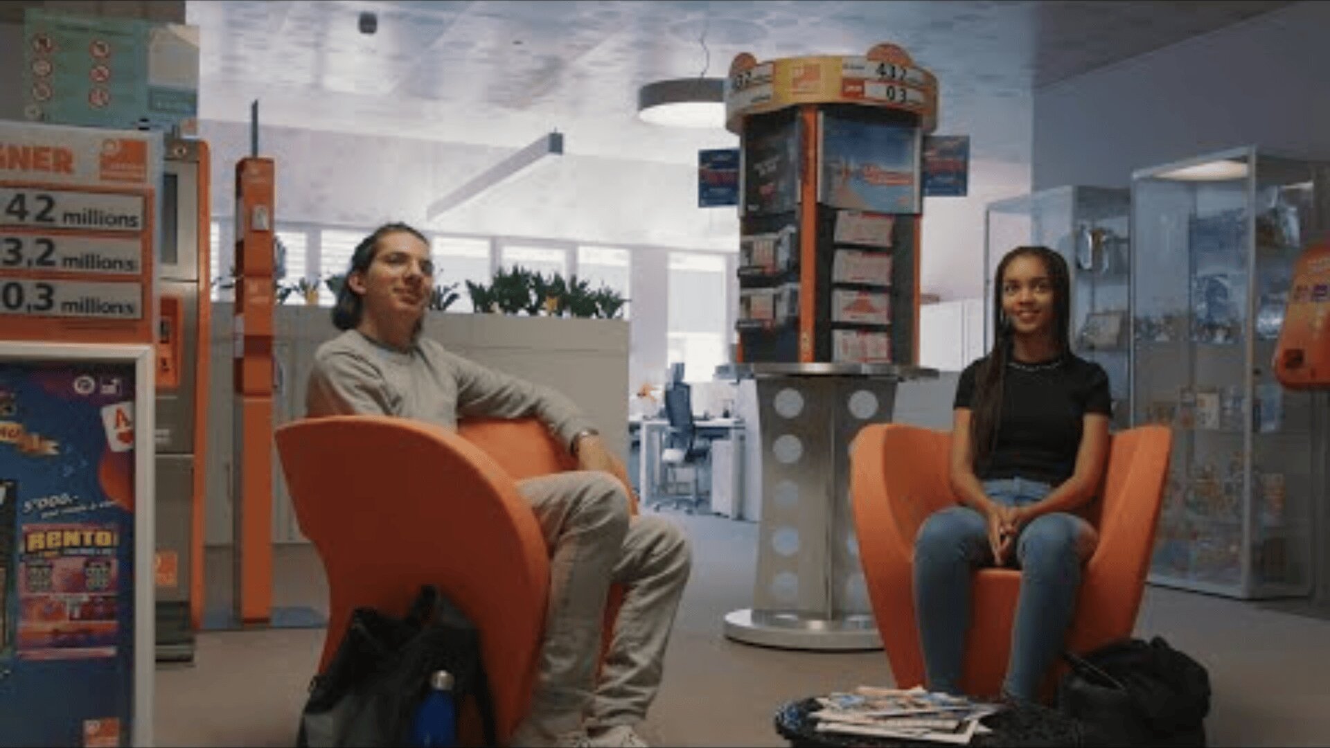 Deux personnes sont assises sur des chaises orange vif dans un bureau moderne ou une salle d'attente, avec des étagères, des brochures et un présentoir de billets de loterie en arrière-plan. Elles semblent toutes deux détendues et sont tournées vers l'avant.