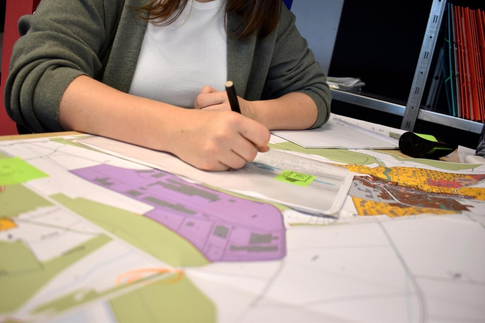 Une personne aux cheveux bruns, vêtue d'une chemise blanche et d'un pull-over vert, dessine ou prend des notes sur une grande carte ou un plan d'architecture avec des zones colorées, à l'aide d'un crayon, à un bureau.