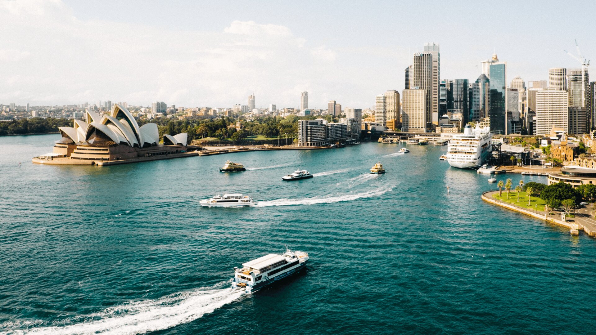 Vue aérienne du port de Sydney avec des bateaux sur l'eau, l'opéra de Sydney à gauche, et la ligne d'horizon de la ville avec de grands bâtiments en arrière-plan, sous un ciel partiellement nuageux.