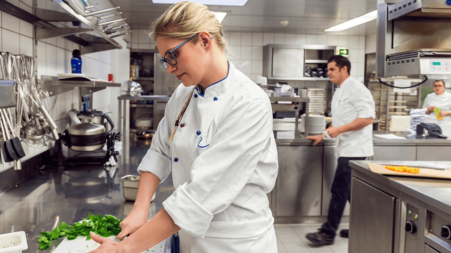 Un chef portant des lunettes et un uniforme blanc prépare des aliments à un comptoir dans une cuisine professionnelle, tandis qu'un autre chef travaille à l'arrière-plan.
