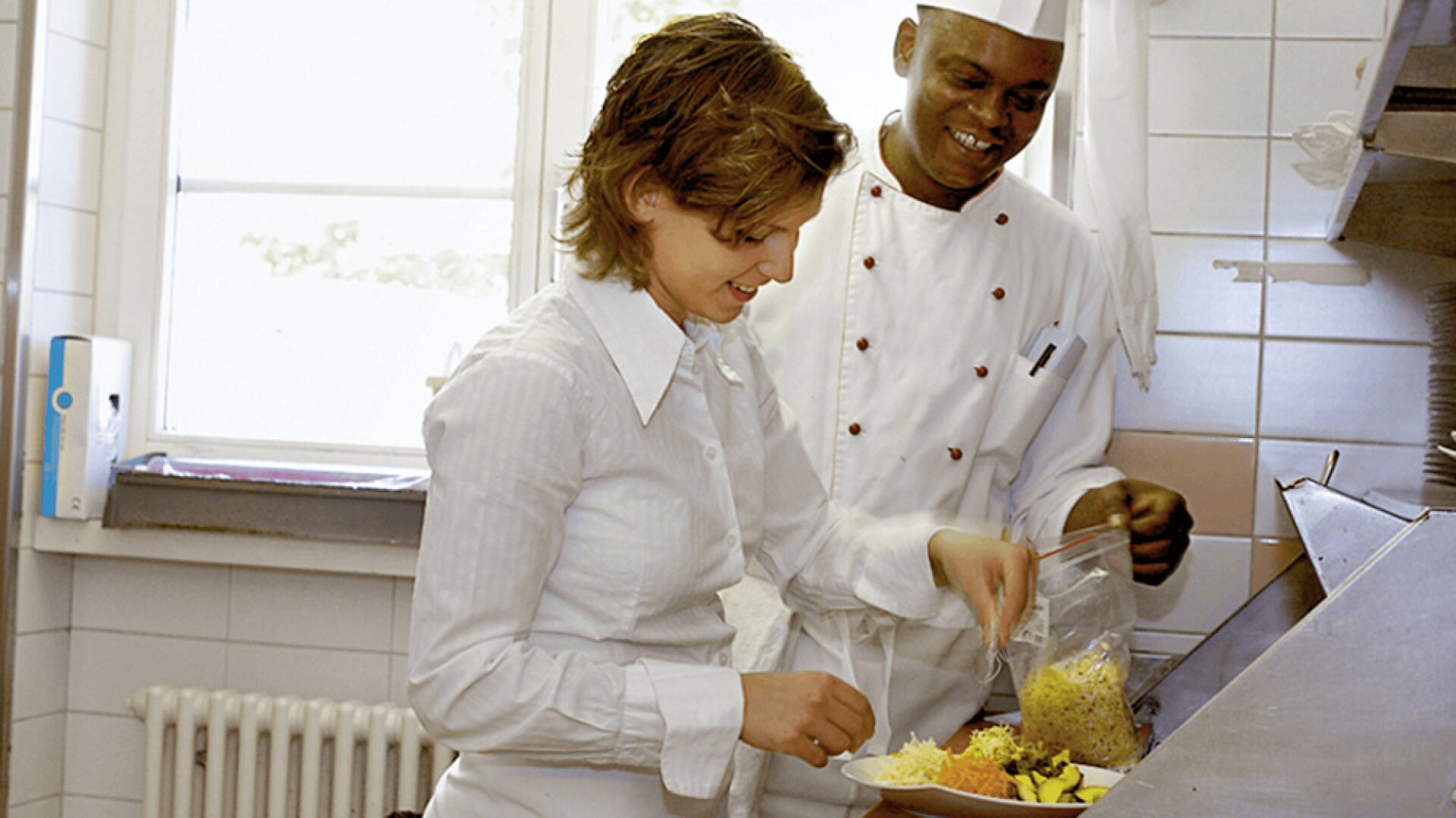 Une femme prépare des aliments à côté d'un chef souriant en uniforme blanc à l'intérieur d'une cuisine, plaçant du fromage râpé et des légumes en tranches sur un comptoir.
