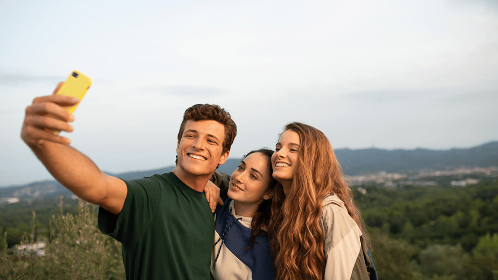 Trois jeunes adultes sourient et posent pour un selfie en plein air avec des arbres verts et des collines lointaines en arrière-plan sous un ciel clair.