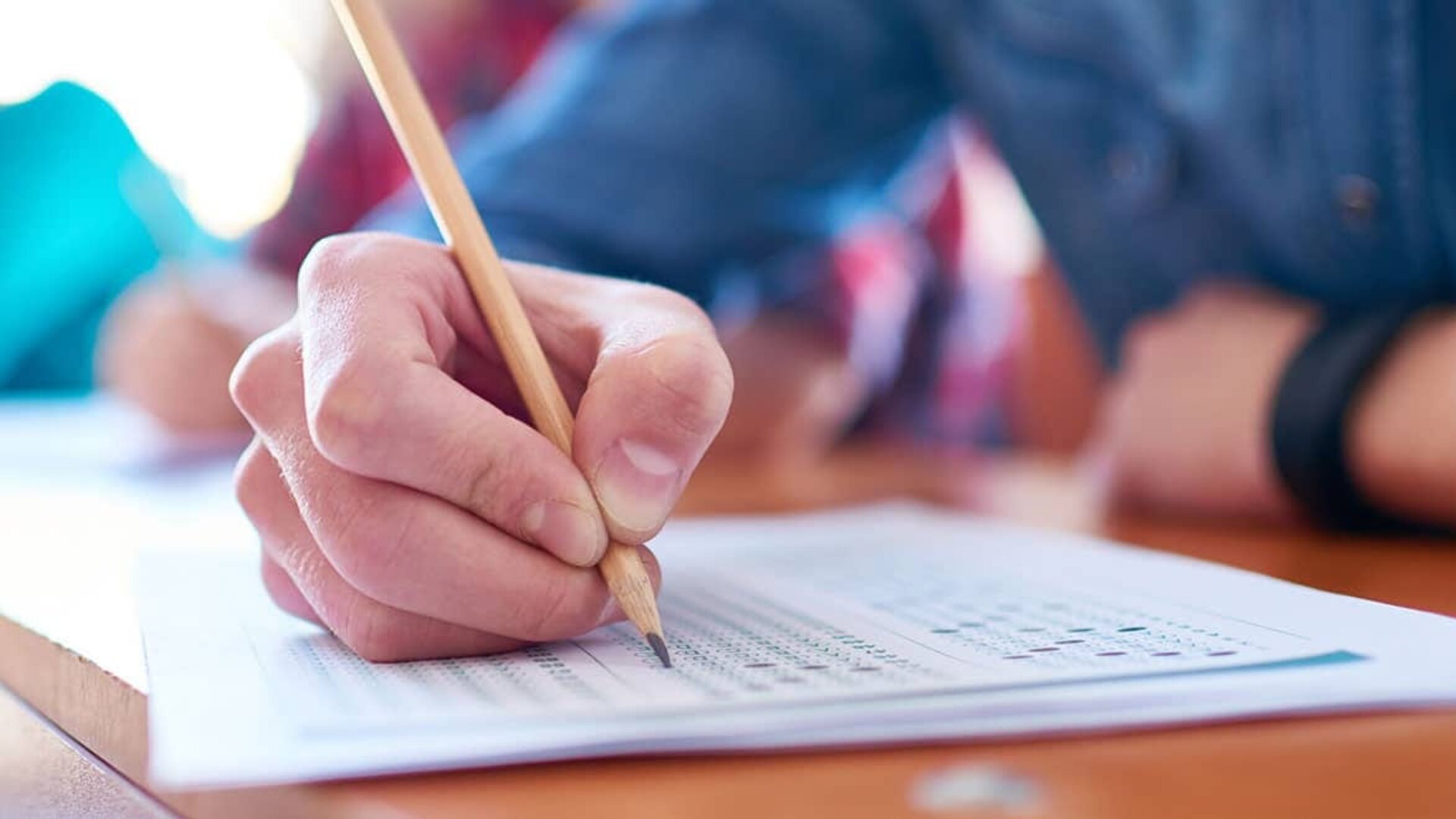 Gros plan de la main d'une personne tenant un crayon et remplissant des réponses sur une feuille de test à choix multiples, avec d'autres étudiants floutés en arrière-plan.