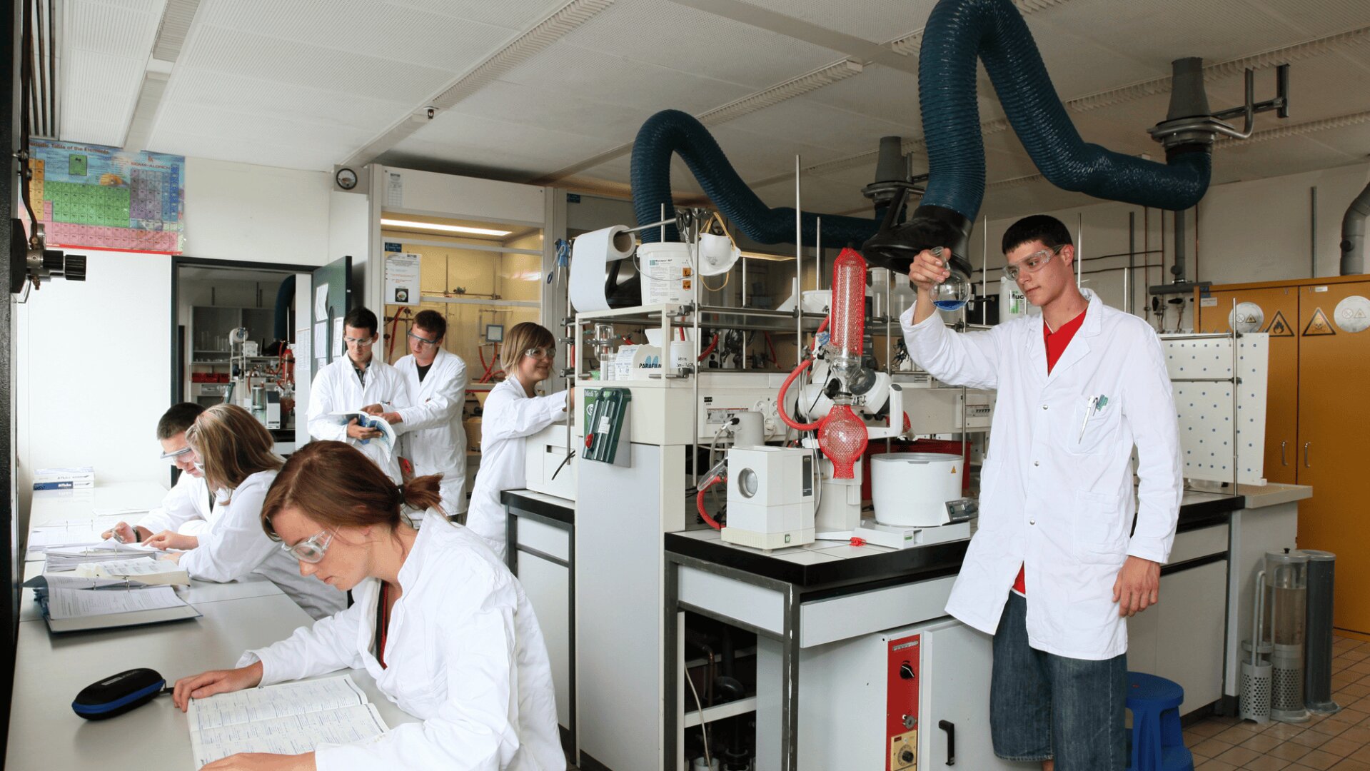 Plusieurs personnes vêtues de blouses blanches travaillent dans un laboratoire très actif avec du matériel scientifique, de la verrerie et des ordinateurs. Une personne se tient au premier plan, tenant une fiole, tandis que d'autres interagissent ou prennent des notes.