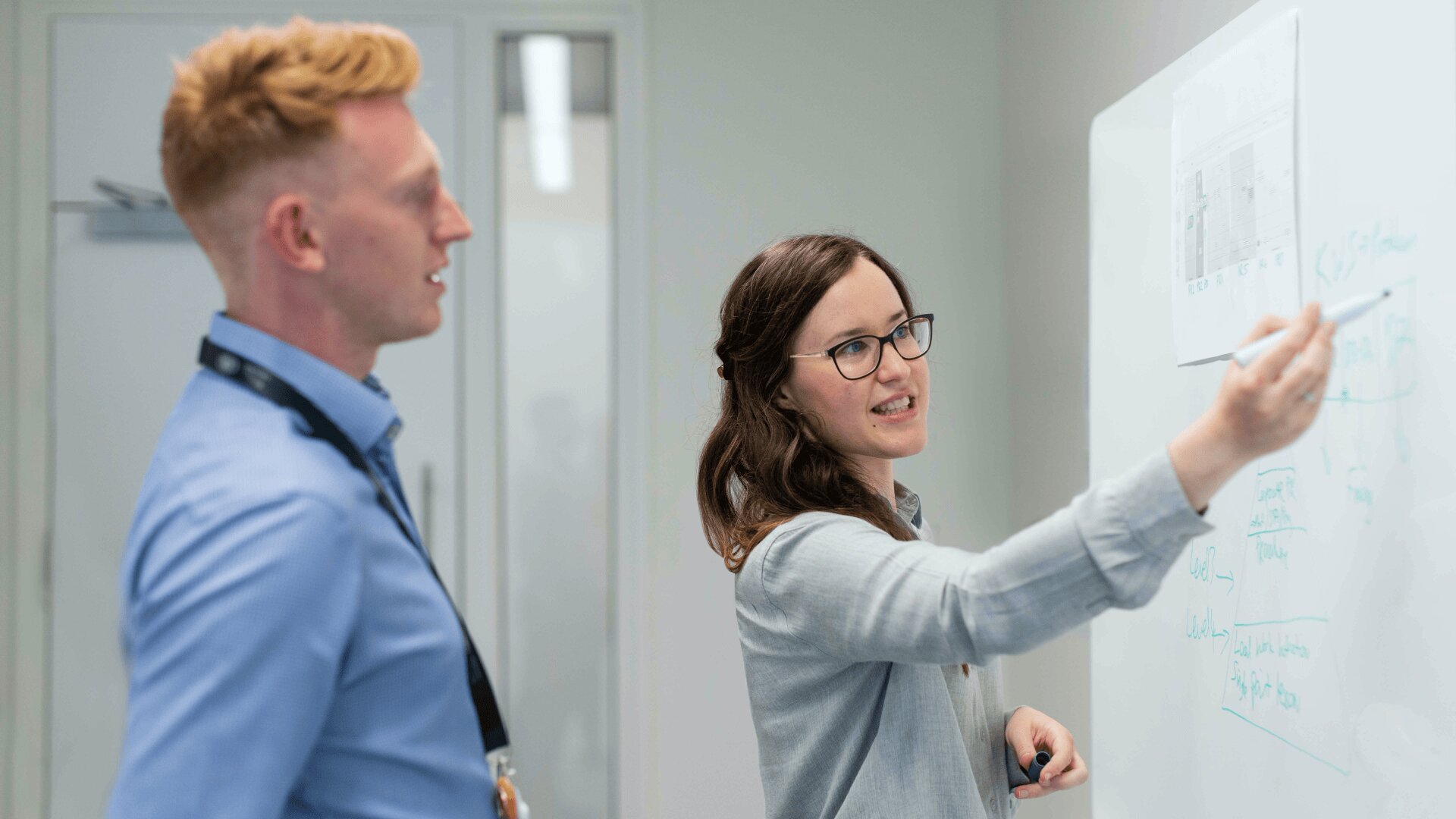 Une femme montre des diagrammes sur un tableau blanc tout en expliquant quelque chose à un homme debout à côté d'elle dans un bureau.