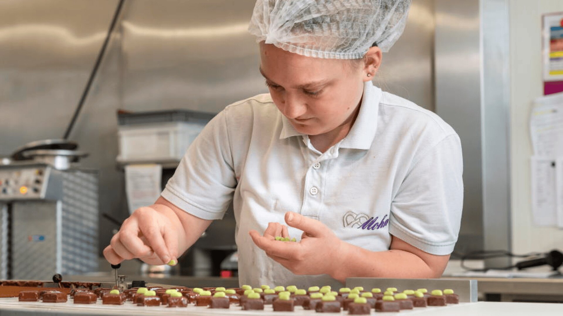 Une personne vêtue d'un polo blanc et d'un filet à cheveux décore soigneusement des rangées de chocolats avec des garnitures vertes dans une cuisine professionnelle.