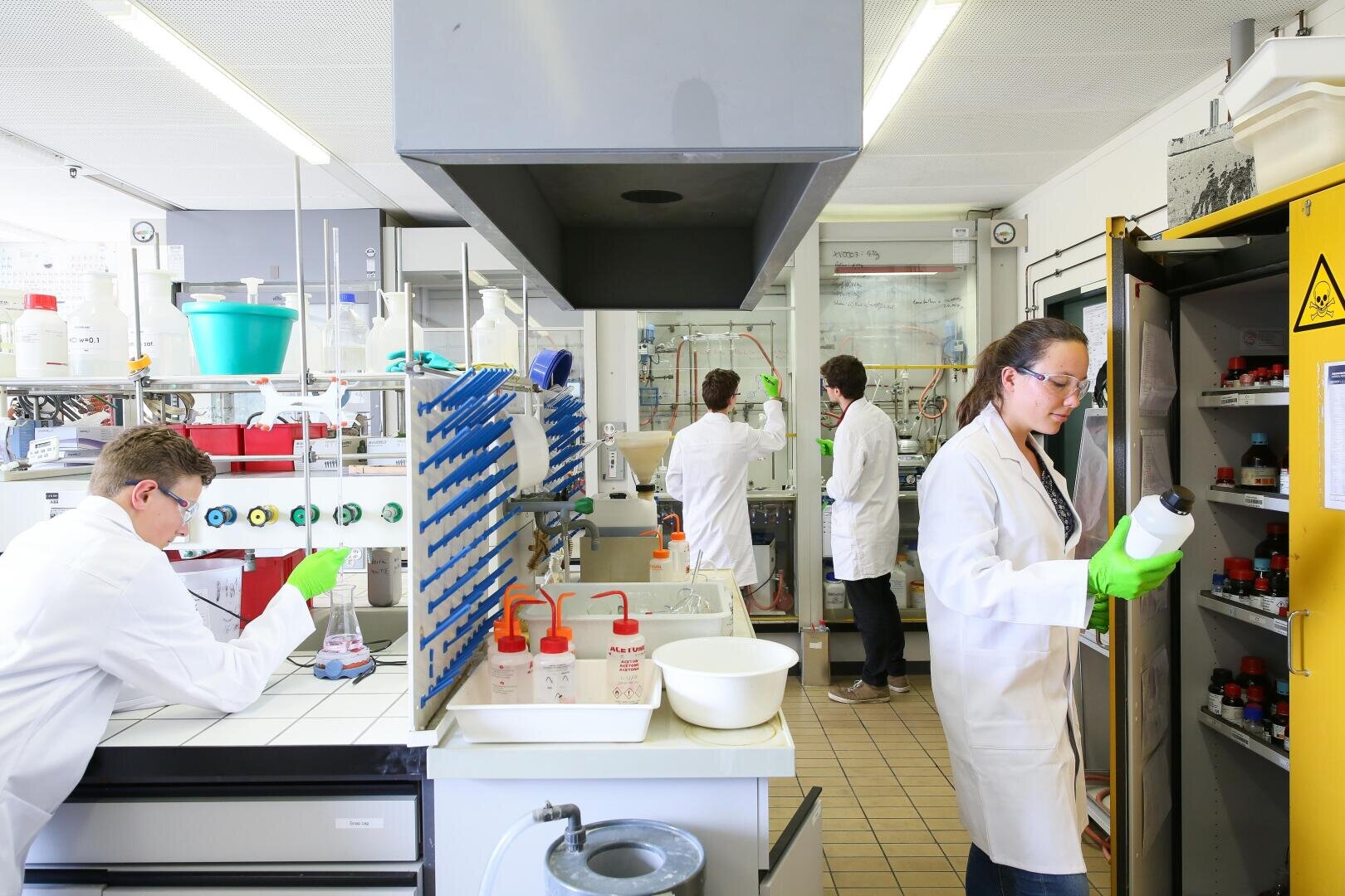 Quatre scientifiques en blouse travaillent dans un laboratoire lumineux ; l'un examine des flacons dans une armoire de stockage de produits chimiques, deux travaillent sur des paillasses et un autre utilise un équipement situé à l'arrière. Les équipements de sécurité et les fournitures de laboratoire sont visibles.