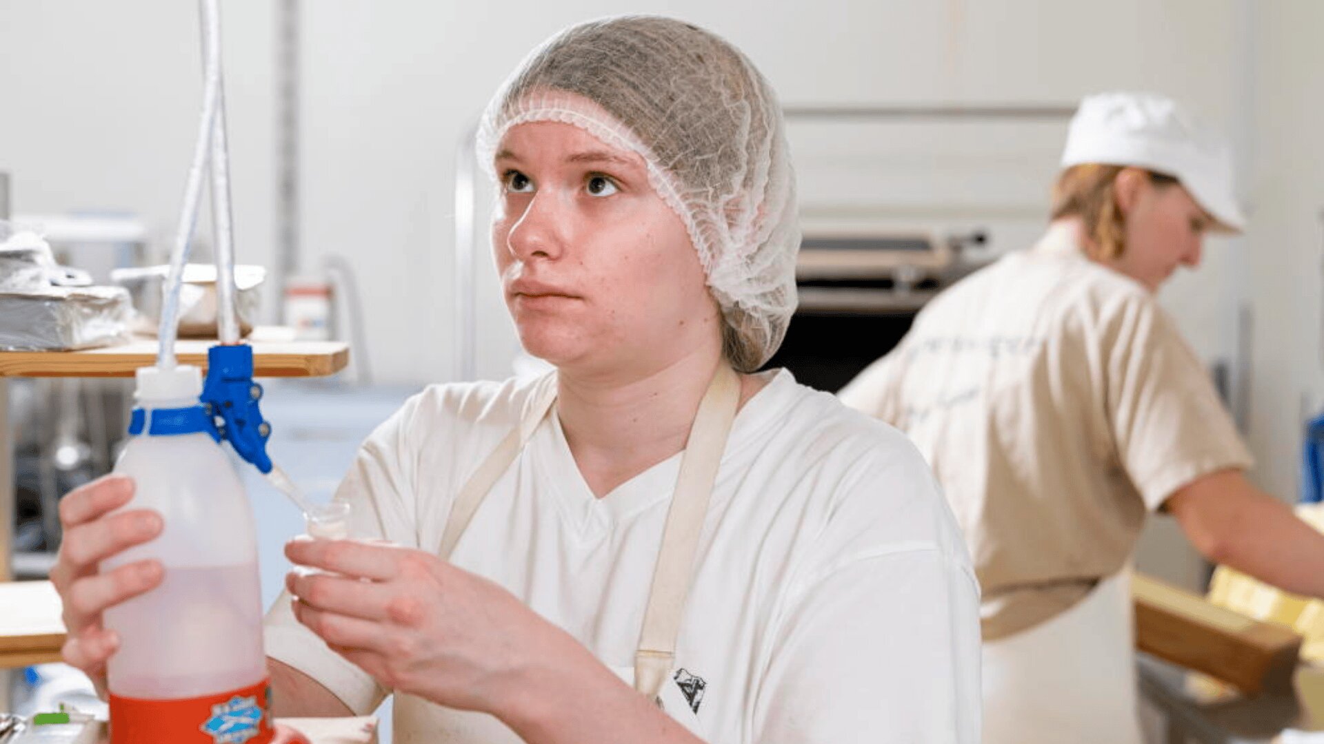 Une personne portant une charlotte, un tablier et une chemise blanche verse un liquide d'un récipient en plastique dans un petit gobelet dans une cuisine ou une zone de préparation des aliments. Une autre personne travaille à l'arrière-plan.