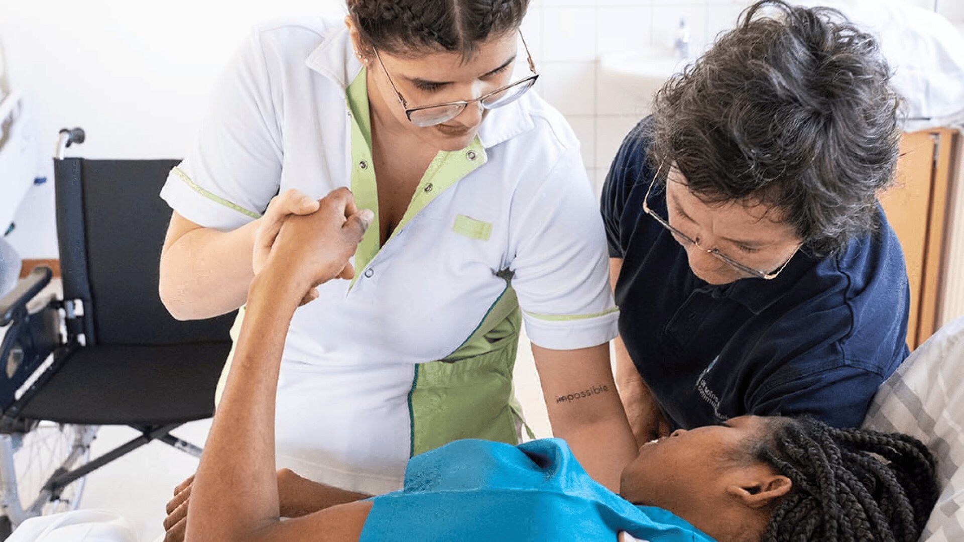 Deux professionnels de la santé aident une femme allongée dans un lit d'hôpital, en lui tenant le bras et en la soutenant. La femme semble recevoir des soins ou une assistance physique. Un fauteuil roulant est visible à l'arrière-plan.