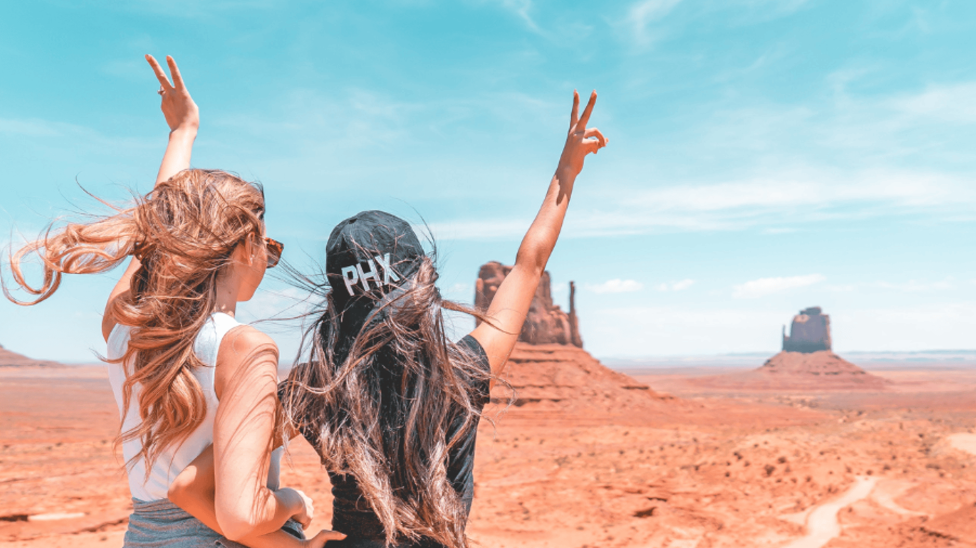 Deux femmes aux cheveux longs et aux lunettes de soleil, l'une portant une casquette PHX noire, se tiennent dans un paysage désertique aux formations rocheuses rouges, levant les mains en signe de paix sous un ciel d'un bleu éclatant.