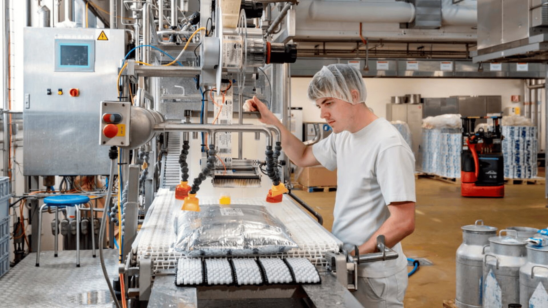 Un ouvrier en uniforme blanc et coiffé d'une charlotte surveille une machine d'emballage dans une usine. Des sacs scellés se déplacent sur un tapis roulant entouré d'équipements industriels et de machines à l'arrière-plan.