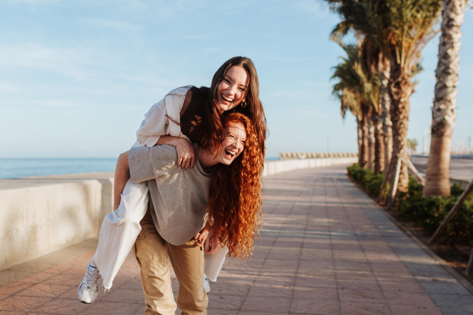 Une jeune femme joyeuse aux longs cheveux bruns se fait monter sur le dos d'un jeune homme aux cheveux roux sur une promenade ensoleillée en bord de mer, bordée de palmiers. Tous deux rient et profitent de l'instant présent.