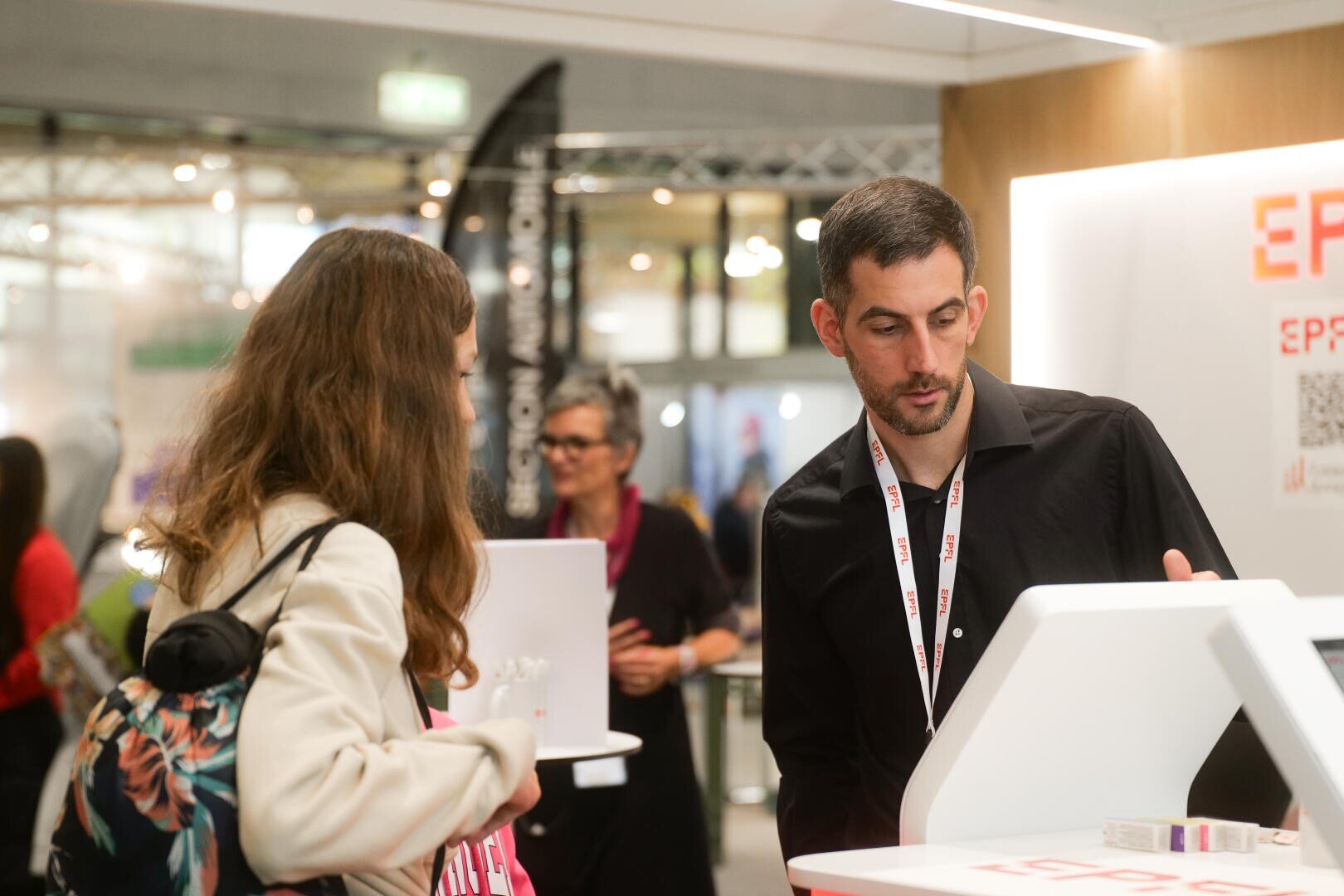 Un homme vêtu d'une chemise noire discute avec une femme aux longs cheveux bruns et portant un sac à dos fleuri sur un stand de salon. D'autres personnes et des stands d'exposition sont visibles dans l'arrière-plan flou.