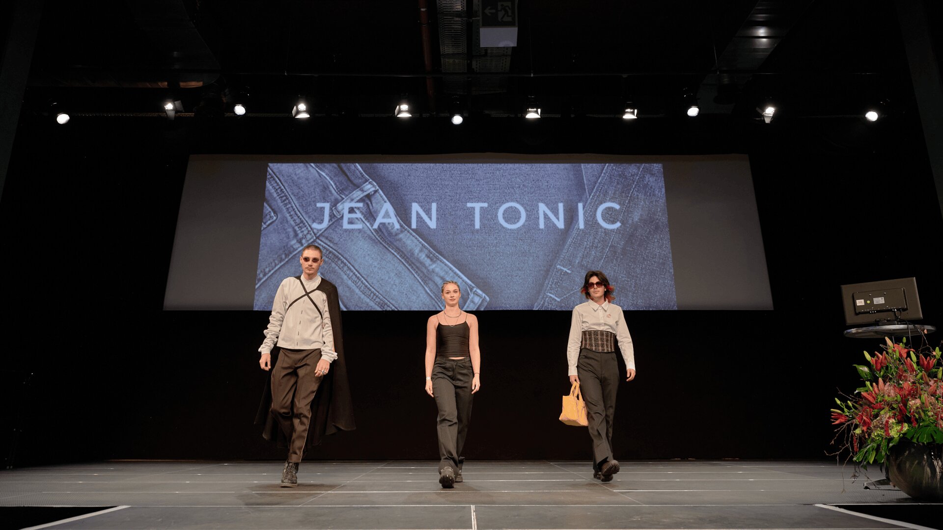Trois mannequins marchent avec assurance sur une piste lors d'un défilé de mode. Derrière elles, un grand écran affiche les mots JEAN TONIC sur un fond de tissu denim. La scène est bien éclairée et décorée de fleurs sur la droite.