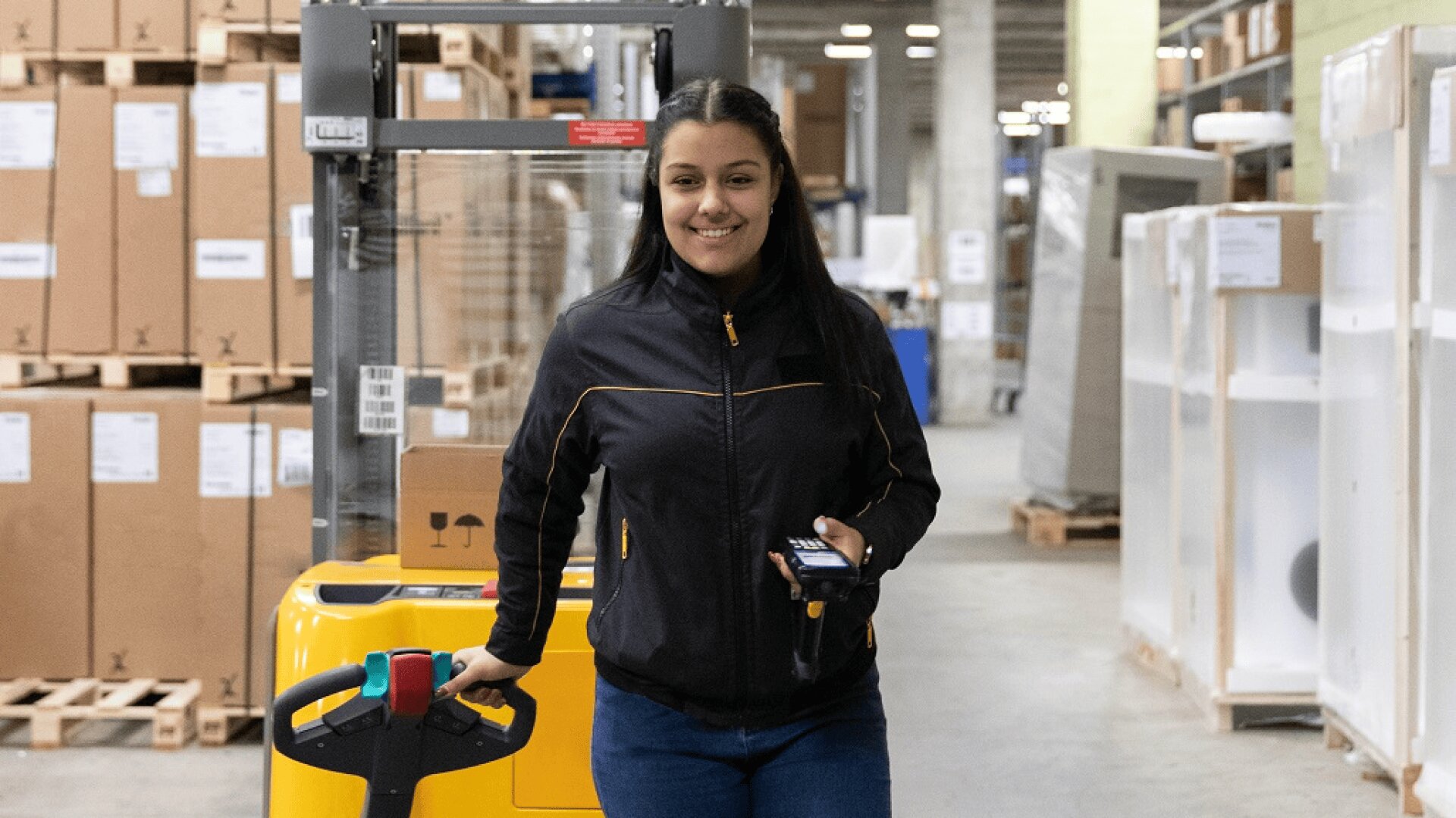 Une femme portant une veste noire se tient dans un entrepôt, souriante et tenant un appareil portable dans une main et la poignée d'un transpalette jaune dans l'autre. Des boîtes et des étagères sont visibles à l'arrière-plan.