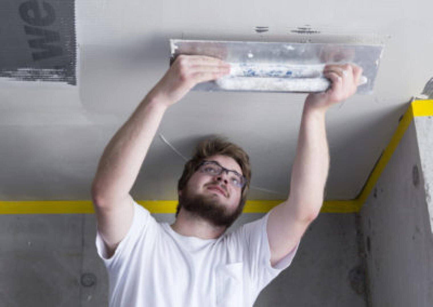 Un homme portant des lunettes et une barbe lisse le plâtre d'un plafond à l'aide d'une truelle. Il porte un tee-shirt blanc et travaille dans une pièce partiellement terminée avec des murs en béton et du ruban adhésif jaune sur les bords.
