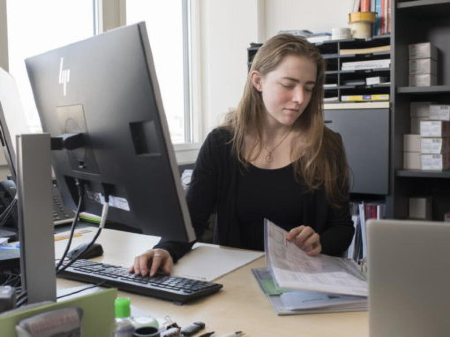 Une femme est assise à un bureau, travaillant sur un ordinateur tout en tenant et en regardant des documents. Des étagères contenant des dossiers et des fournitures de bureau se trouvent à l'arrière-plan.