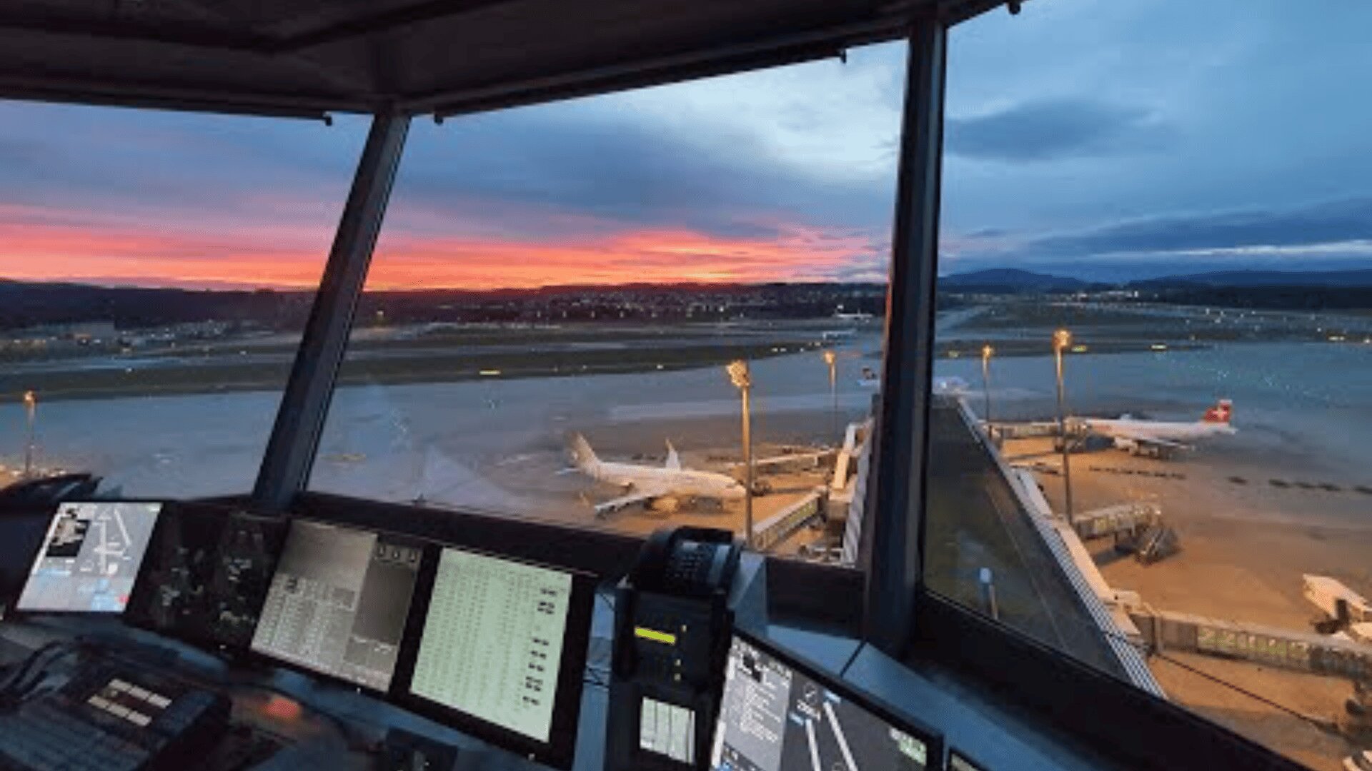 Vue d'une tour de contrôle d'aéroport au coucher du soleil, montrant des panneaux de contrôle au premier plan et des avions stationnés à des portes sur la piste à l'extérieur, sous un ciel coloré.